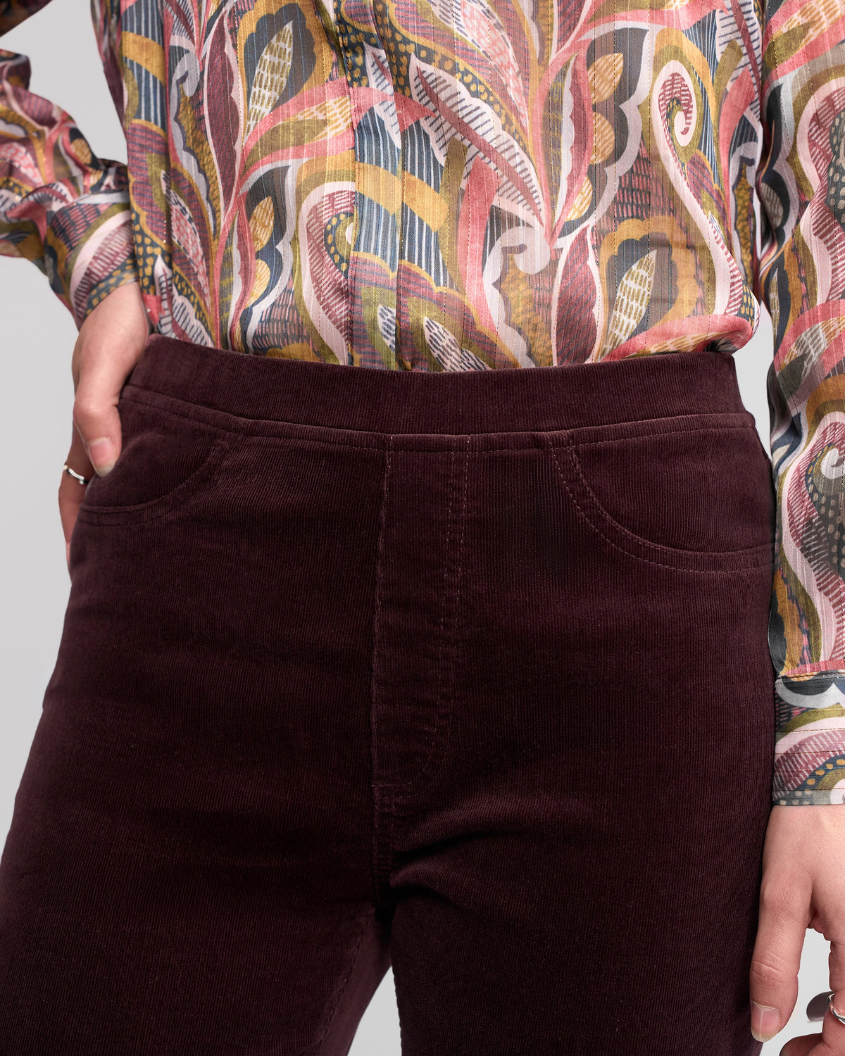 A person is shown from mid-torso to thigh, wearing Vassalli's Bay Cord Pull On pants in Merlot and a colorful, swirling patterned long-sleeve blouse.