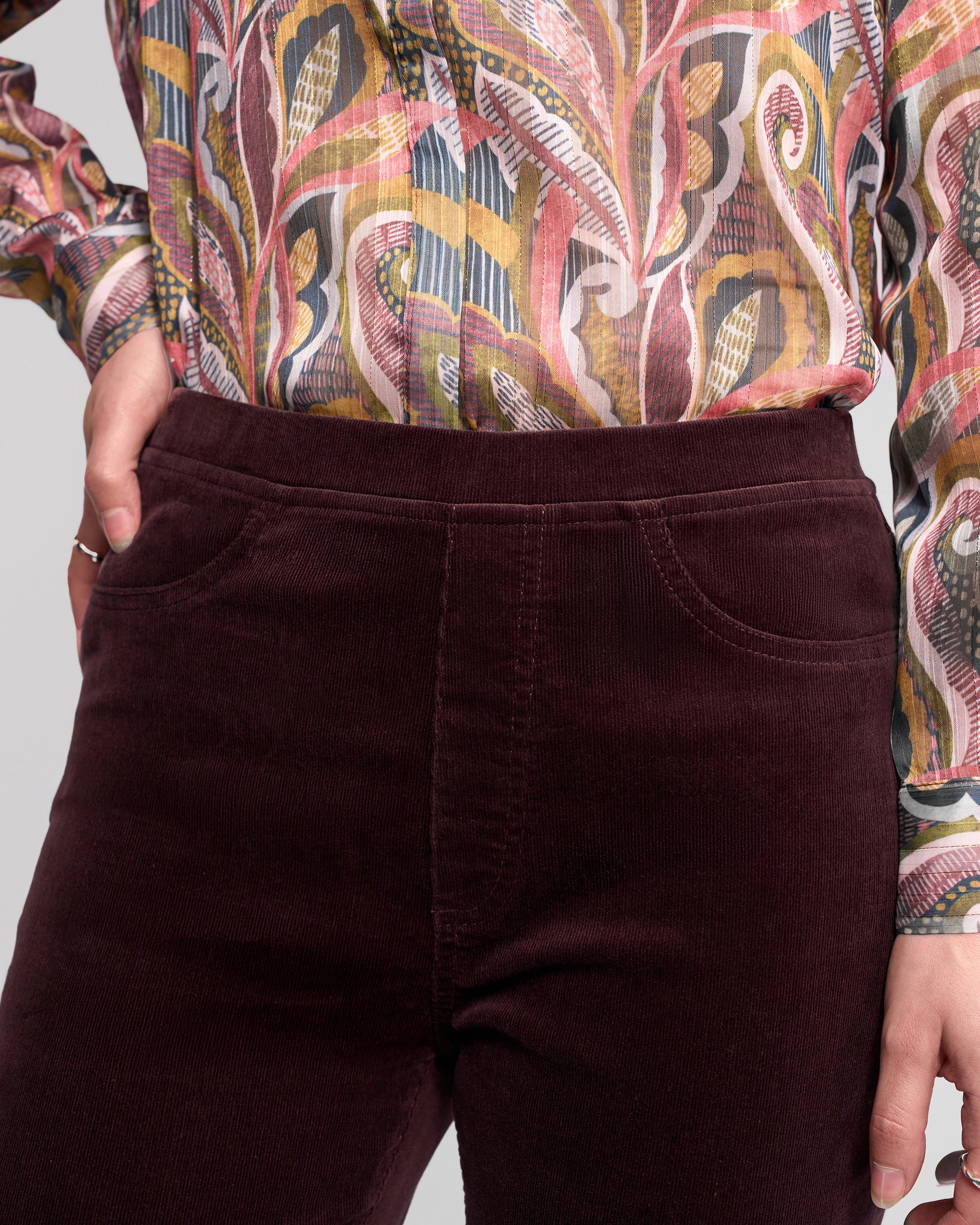 A person is shown from mid-torso to thigh, wearing Vassalli's Bay Cord Pull On pants in Merlot and a colorful, swirling patterned long-sleeve blouse.