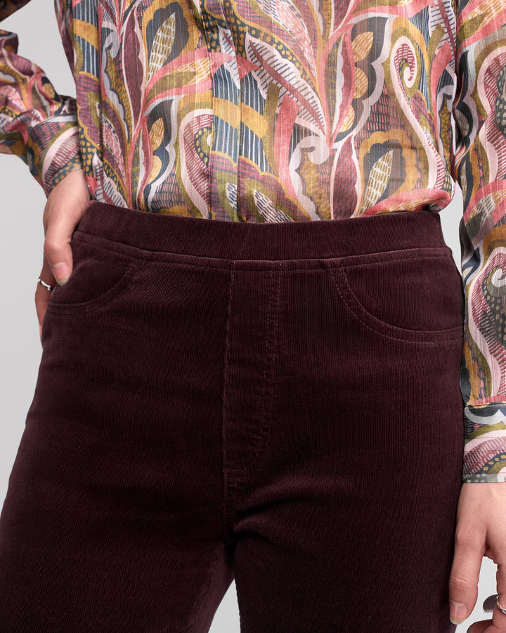 A person is shown from mid-torso to thigh, wearing Vassalli's Bay Cord Pull On pants in Merlot and a colorful, swirling patterned long-sleeve blouse.