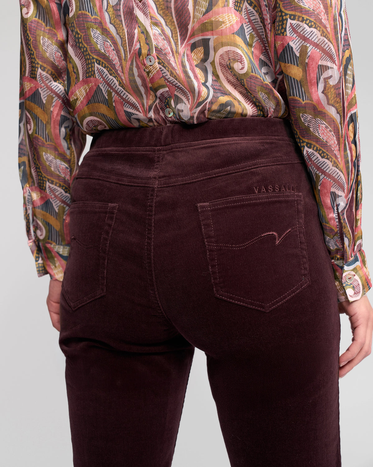 A person is shown from the back wearing a colorful patterned blouse and Vassalli Bay Cord Pull On - Merlot slim-leg pants with pockets, featuring "Vassalli" stitched above the right pocket.