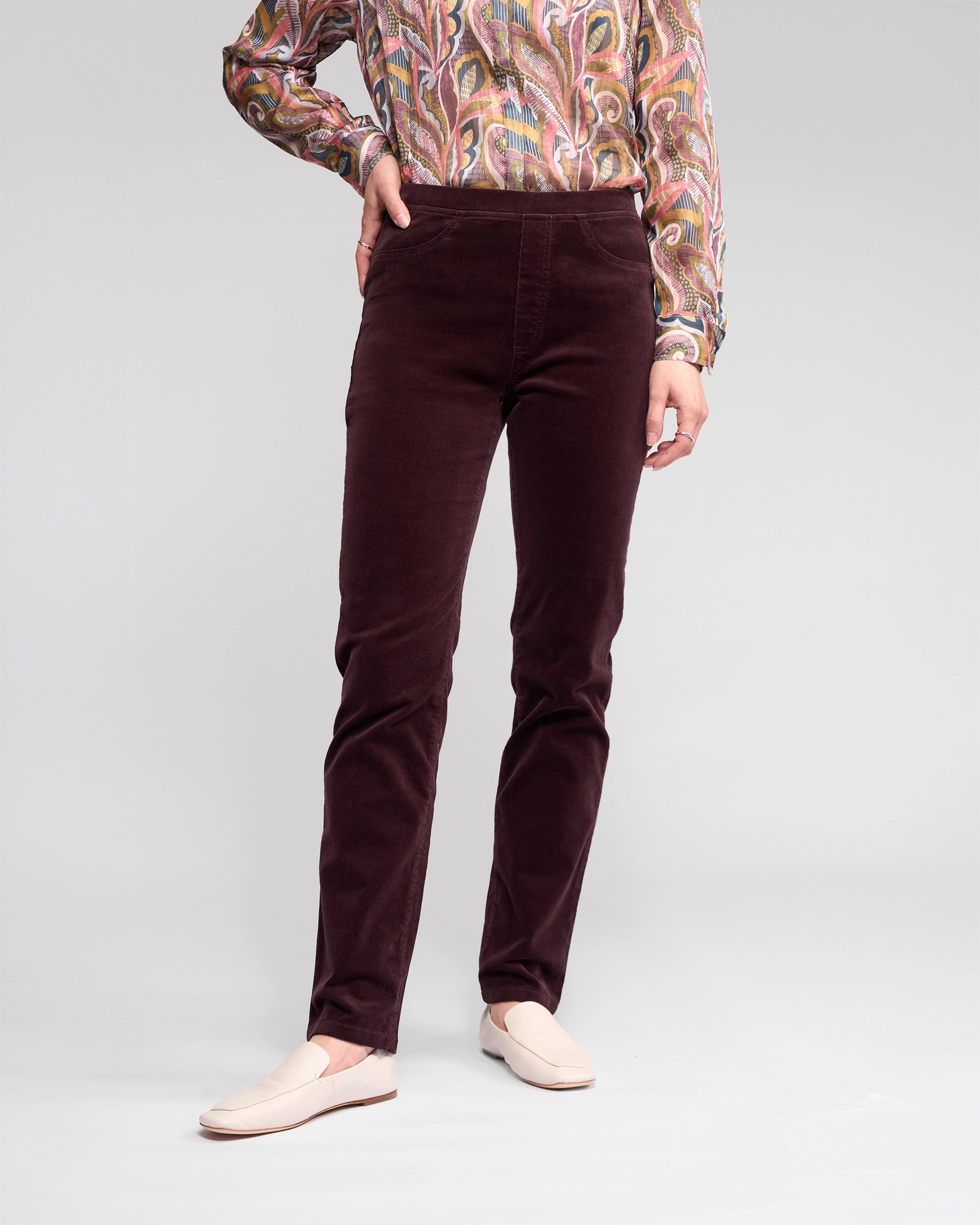 A person wears Vassalli's Bay Cord Pull On pants in Merlot with a paisley long-sleeve shirt and cream loafers, standing with one hand on their hip against a plain light gray background.