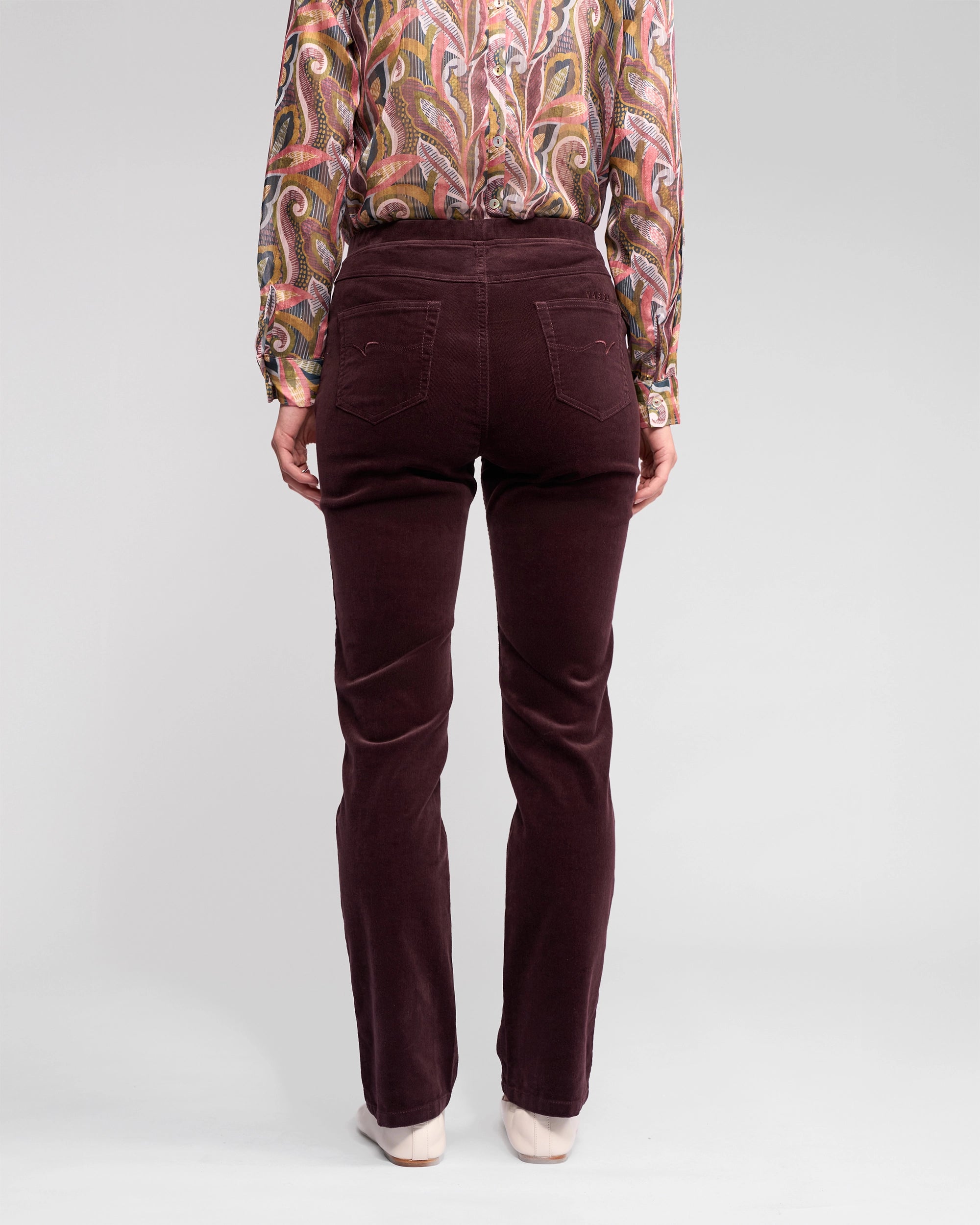 A person stands facing away, wearing Vassalli's Bay Cord Pull On - Merlot slim-leg pant and a long-sleeve patterned paisley blouse against a plain light gray background.