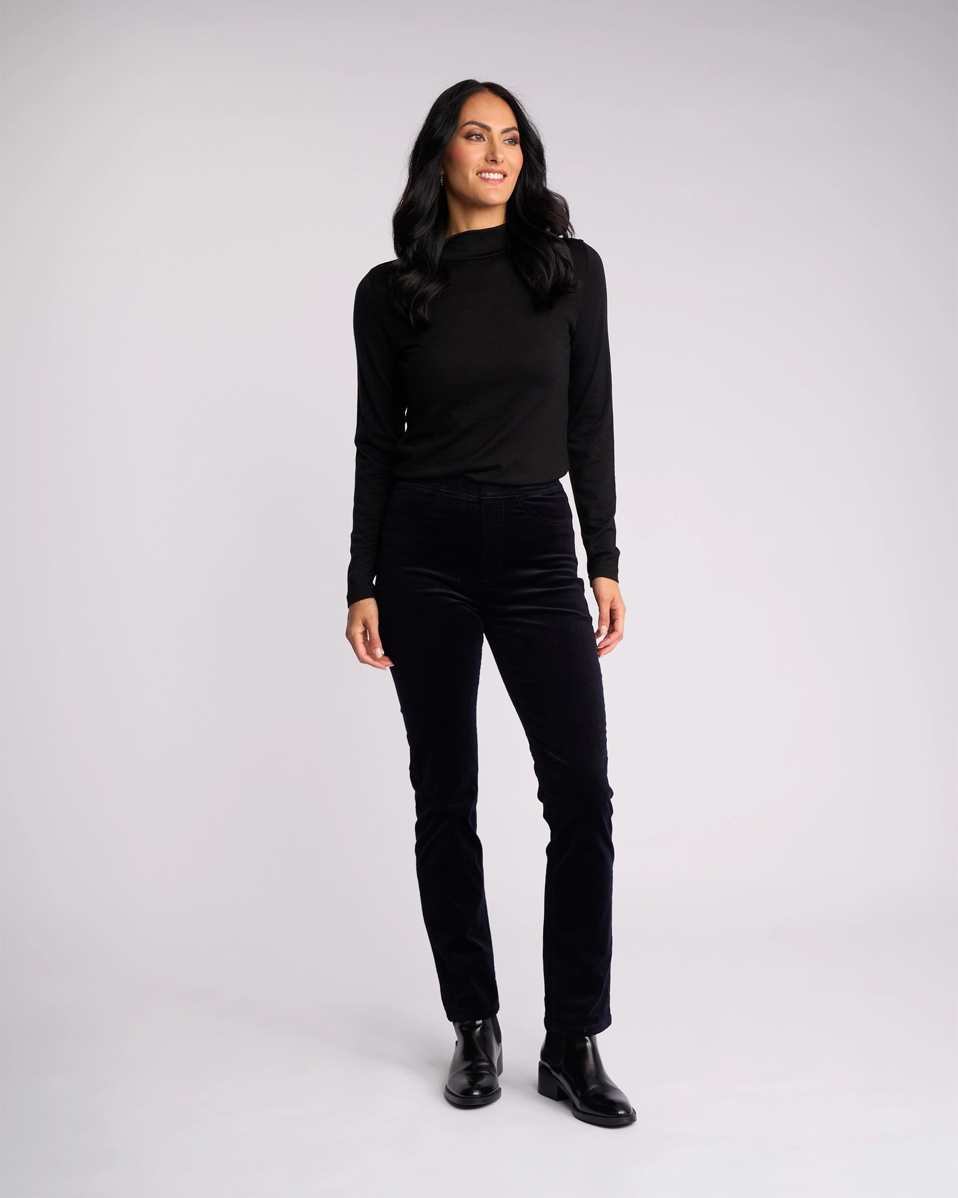 A woman with long dark hair stands against a light background, wearing Vassalli’s Bay Cord Pull On - Midnight pants, a black long-sleeve top, and black ankle boots. She smiles slightly and gazes off to the side.