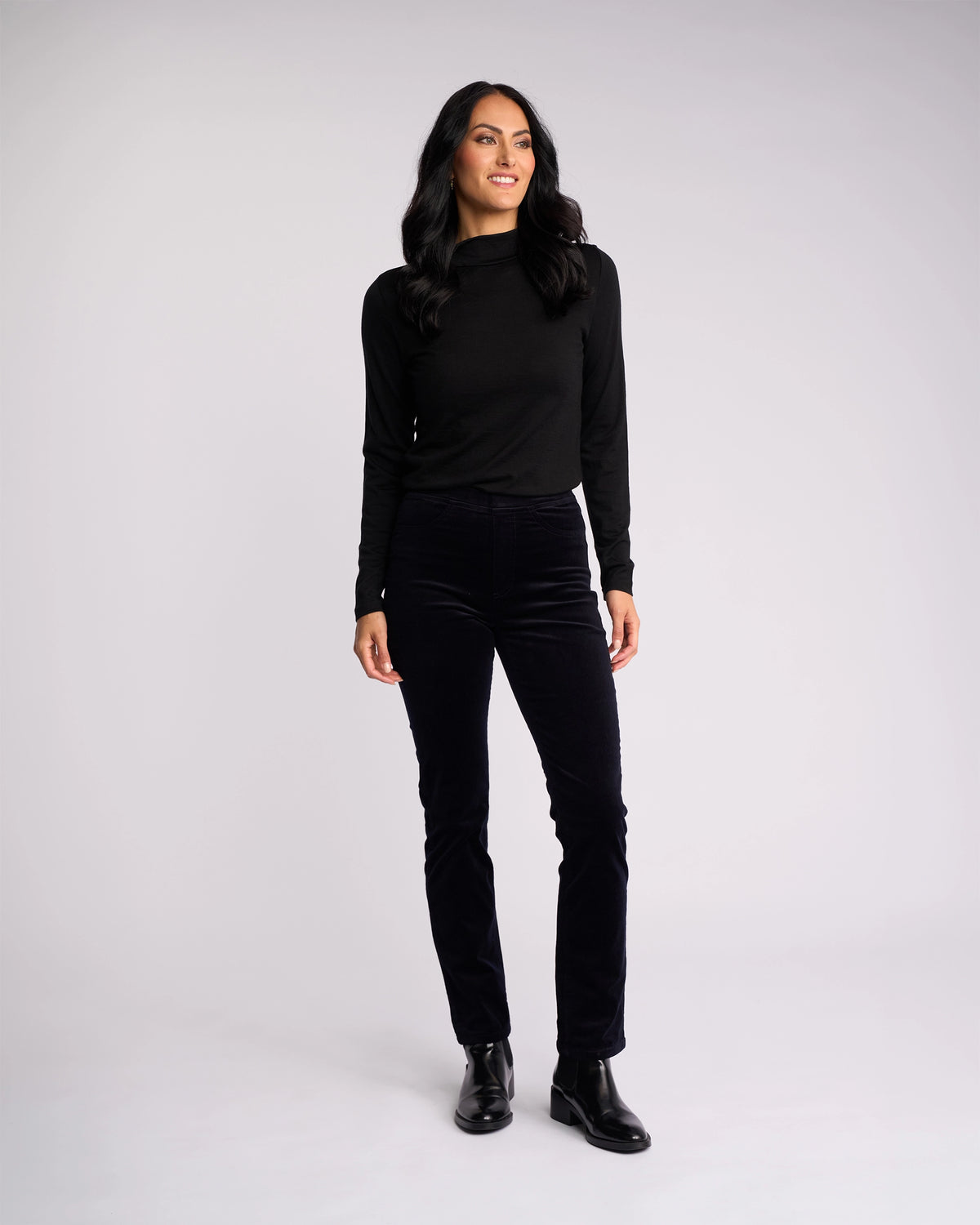 A woman with long dark hair stands against a light background, wearing Vassalli’s Bay Cord Pull On - Midnight pants, a black long-sleeve top, and black ankle boots. She smiles slightly and gazes off to the side.