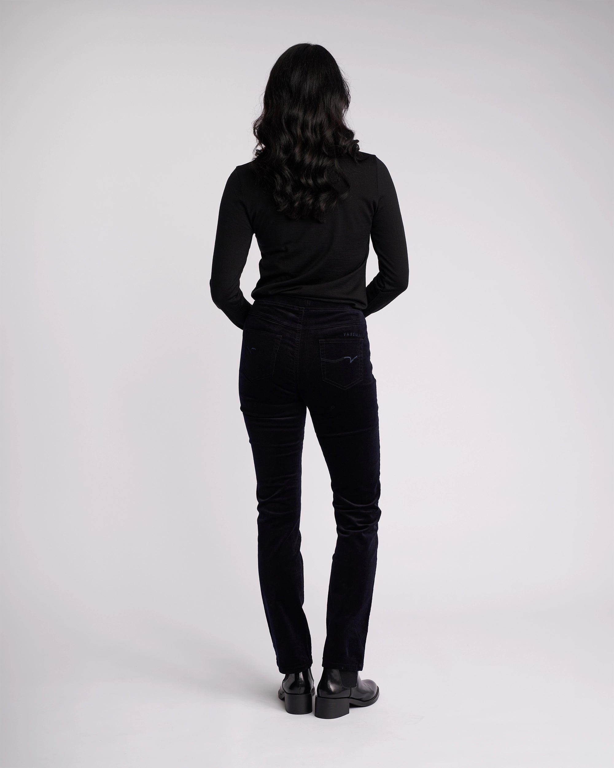 A person with long, wavy dark hair wears a black long-sleeve top and Vassalli Bay Cord Pull On - Midnight slim-leg pants with black shoes, standing facing away from the camera against a plain light background.