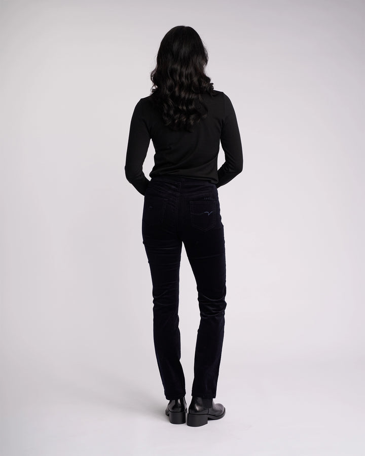 A person with long, wavy dark hair wears a black long-sleeve top and Vassalli Bay Cord Pull On - Midnight slim-leg pants with black shoes, standing facing away from the camera against a plain light background.