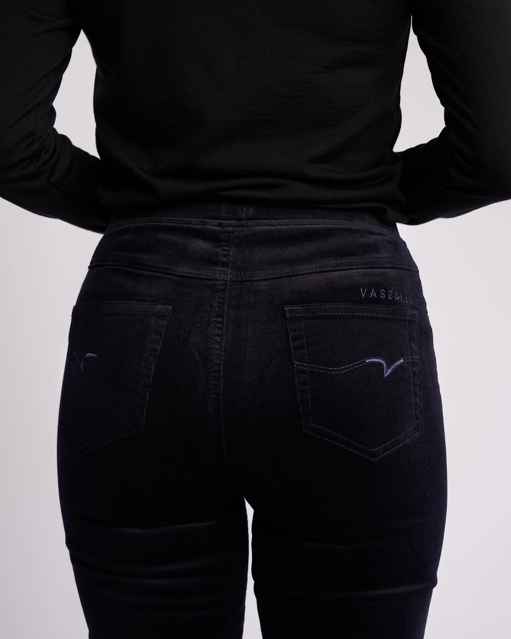 A person wearing Vassalli's Bay Cord Pull On in Midnight is shown from the waist down and back. The slim-leg pants feature stretch pinwale cord fabric, two back pockets with subtle stitching, and "VASSMUI" embroidered above the right pocket.