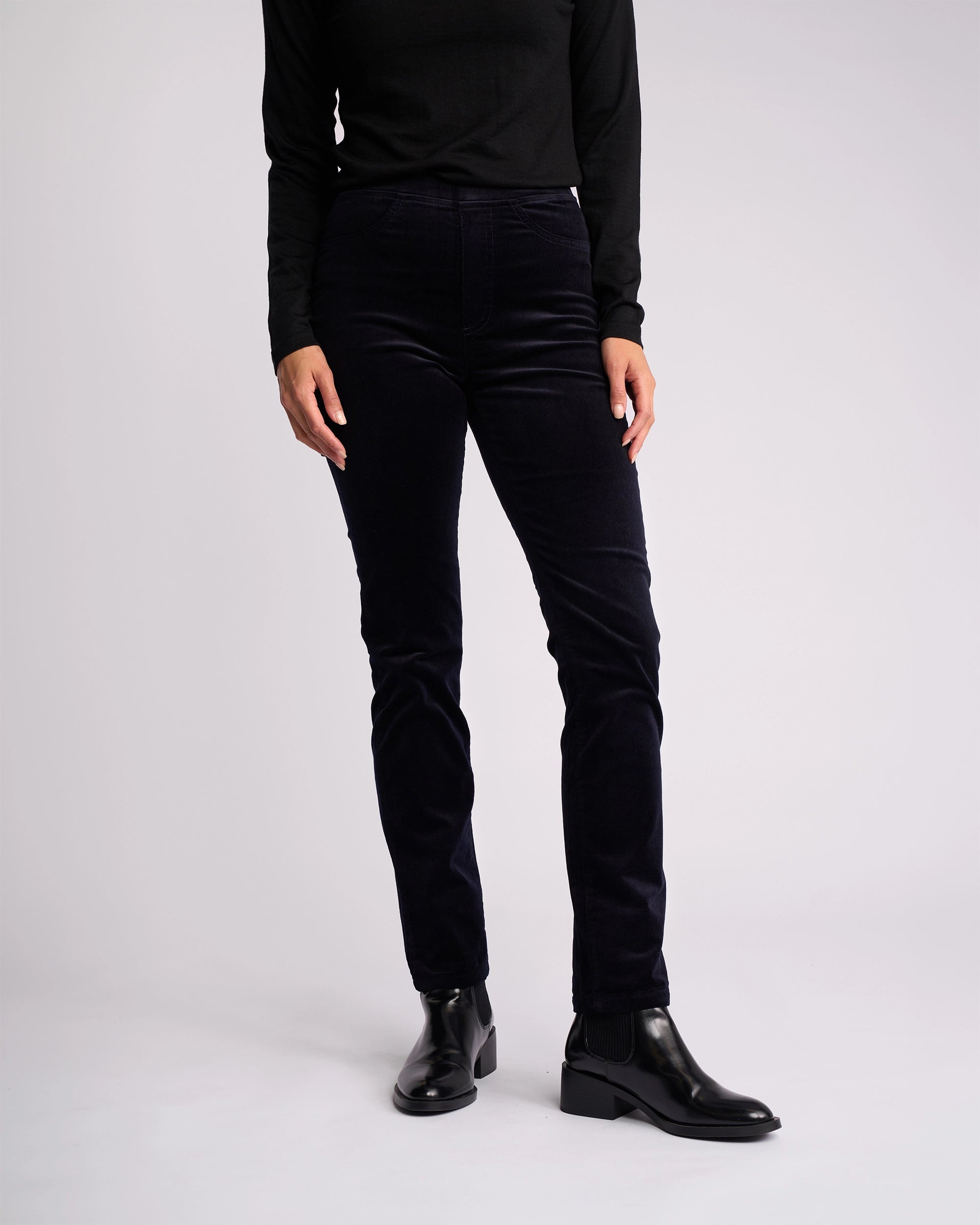 A person stands against a plain background, wearing a long-sleeve black top, Vassalli Bay Cord Pull On - Midnight slim-leg pants, and black ankle boots. Their head is not visible in the image.