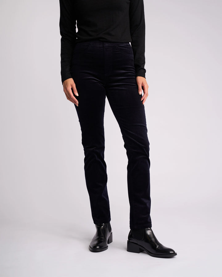 A person stands against a plain background, wearing a long-sleeve black top, Vassalli Bay Cord Pull On - Midnight slim-leg pants, and black ankle boots. Their head is not visible in the image.