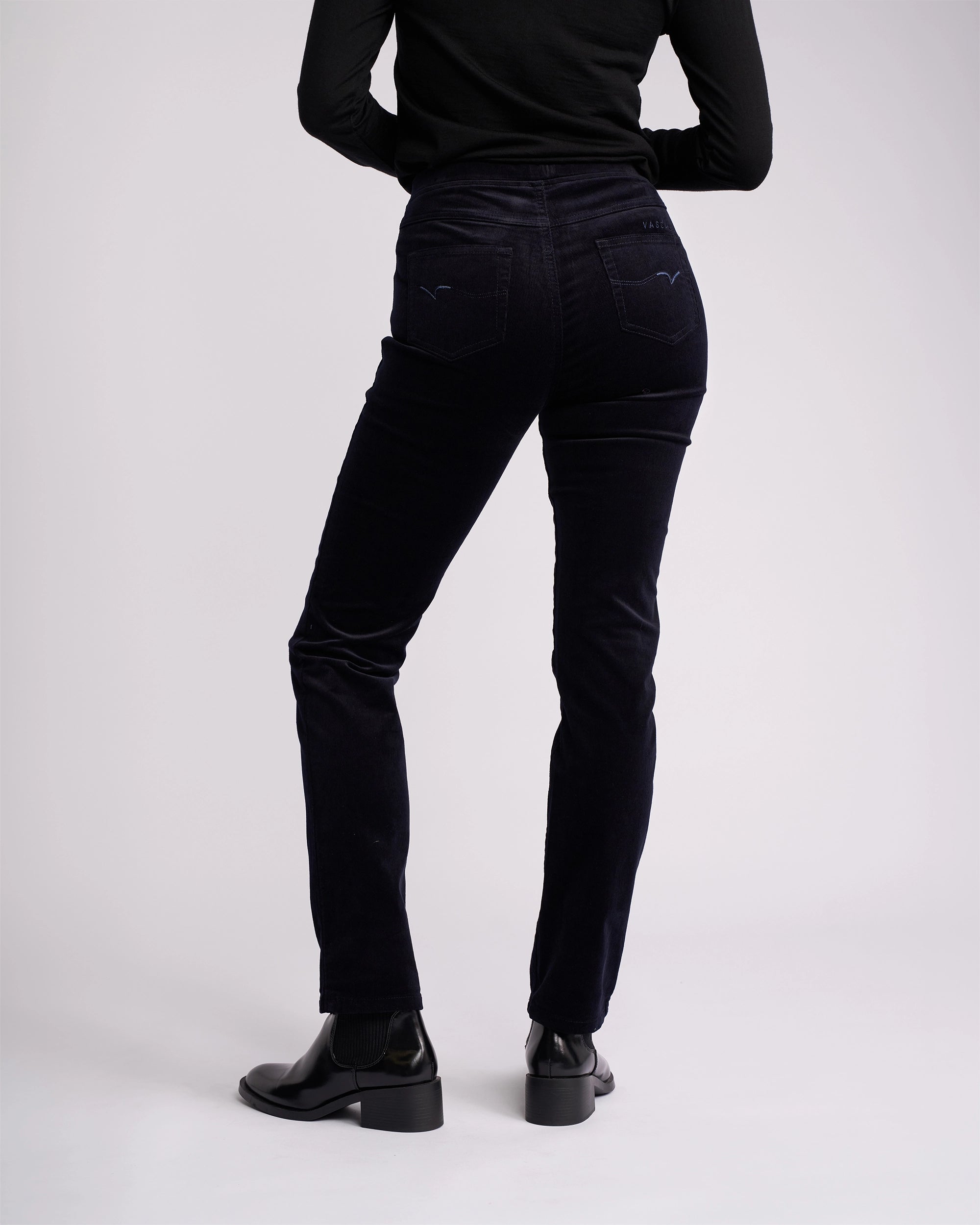 A person, visible from shoulders down, wears a black long-sleeve top, Vassalli Bay Cord Pull On - Midnight pants, and black heeled boots against a plain light background.