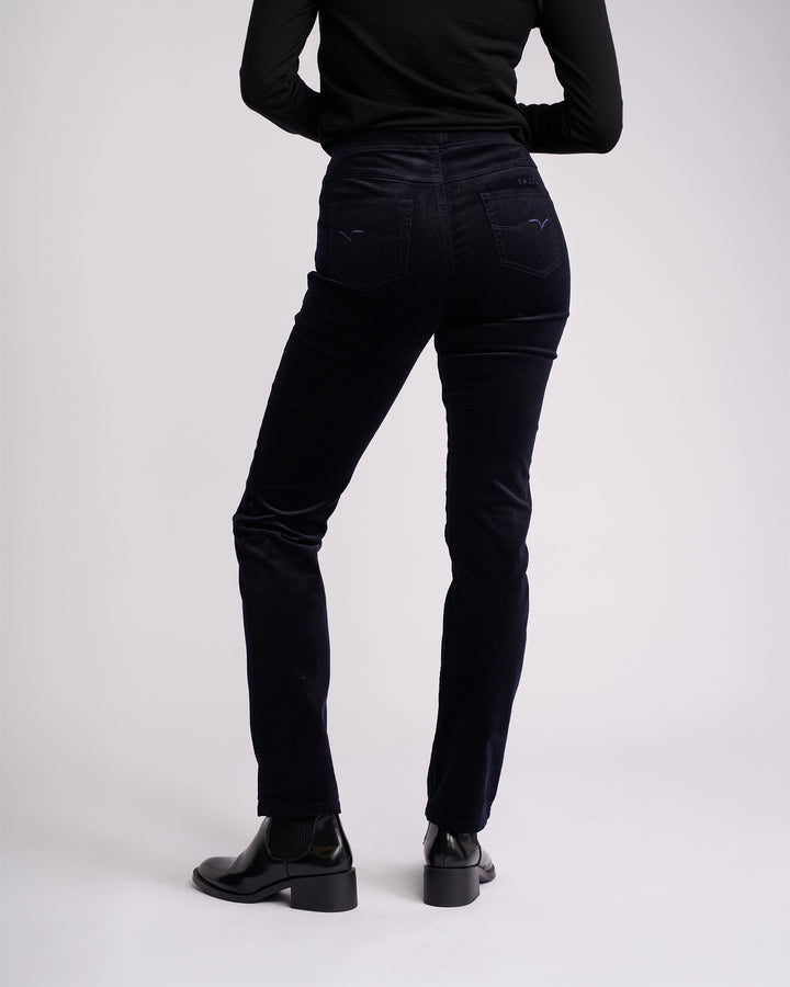 A person, visible from shoulders down, wears a black long-sleeve top, Vassalli Bay Cord Pull On - Midnight pants, and black heeled boots against a plain light background.