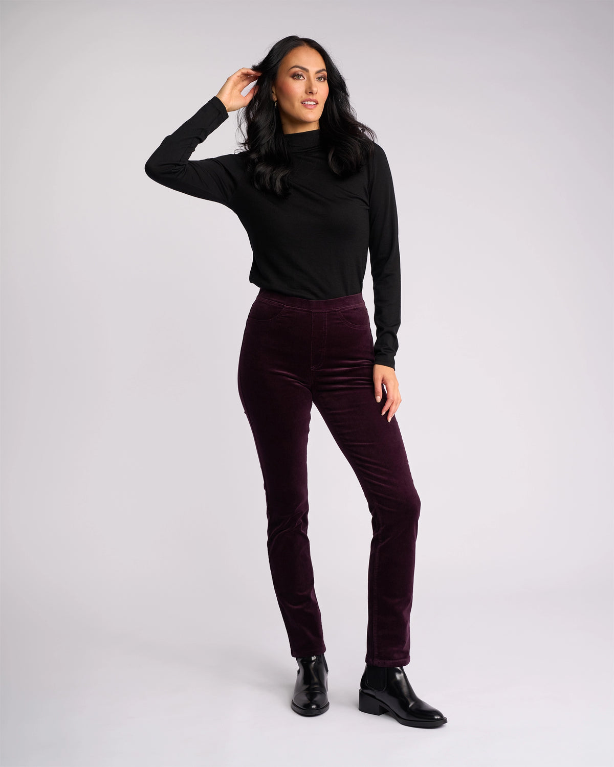 A person stands against a plain white background in a long-sleeve black top, black ankle boots, and Vassalli Bay Cord Pull On pants in Mulberry, with one hand touching their hair and the other resting by their side.