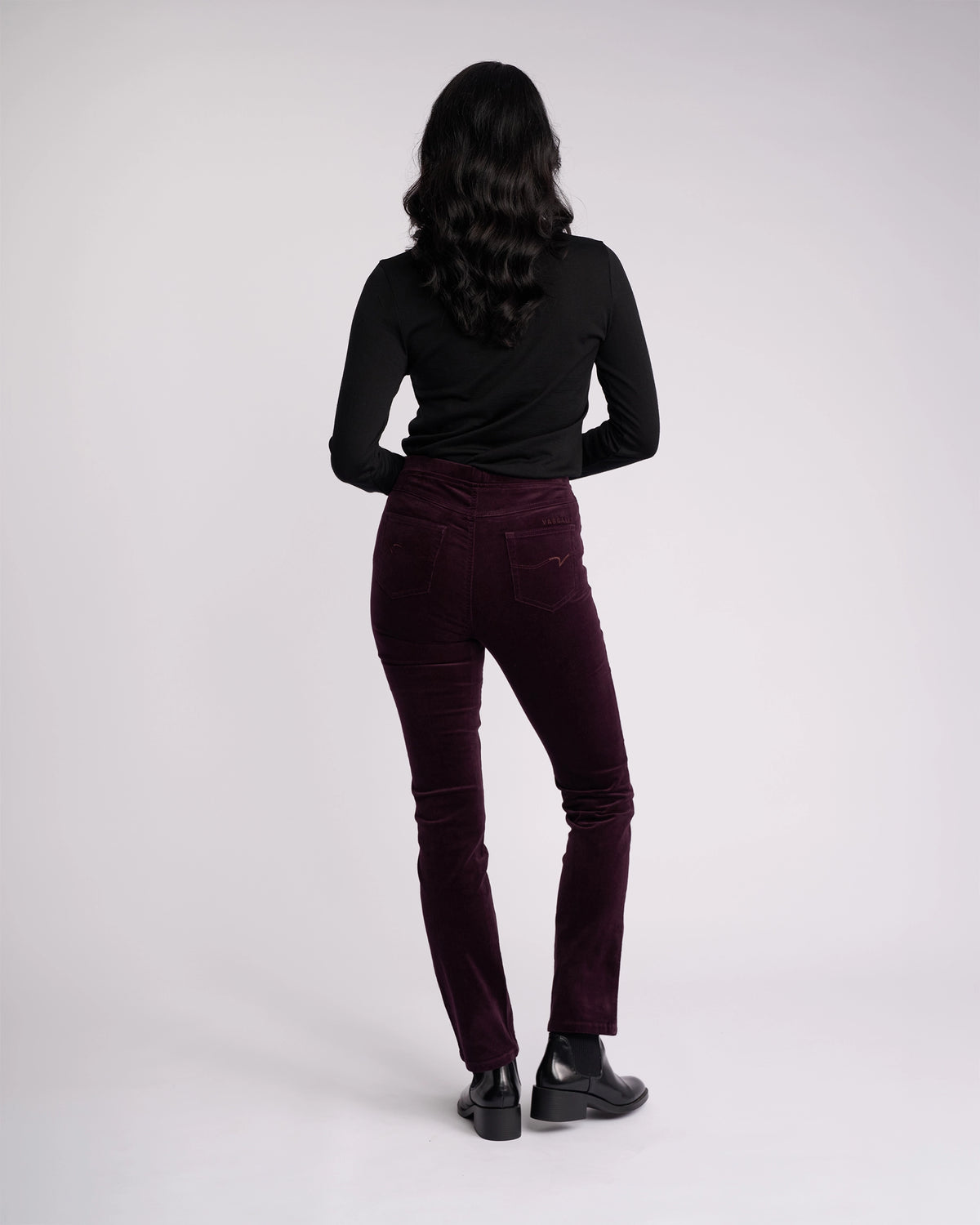 A woman with long wavy hair stands facing away from the camera, wearing a black long-sleeve top, black ankle boots, and Vassalli Bay Cord Pull On pants in Mulberry against a plain light background.