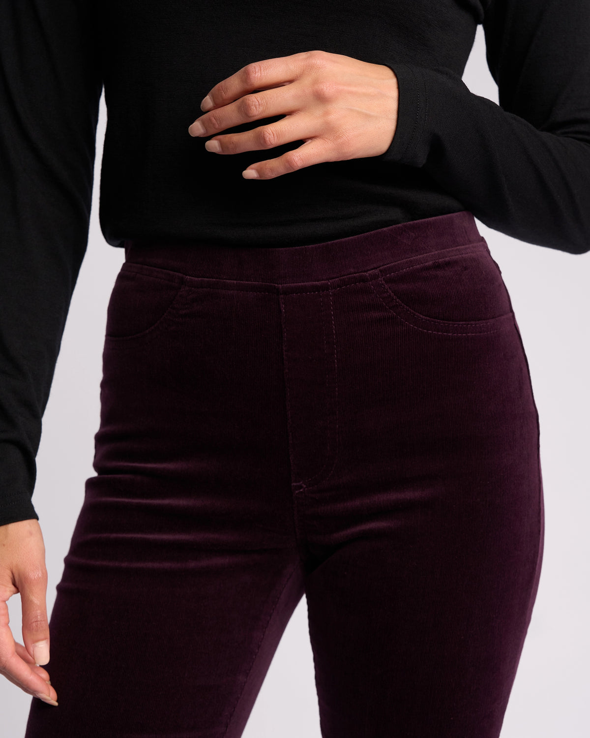 A person is wearing Vassalli’s Bay Cord Pull On pants in Mulberry, paired with a long-sleeved black top. They stand with one hand near their waist; the image is cropped from shoulders to thighs.