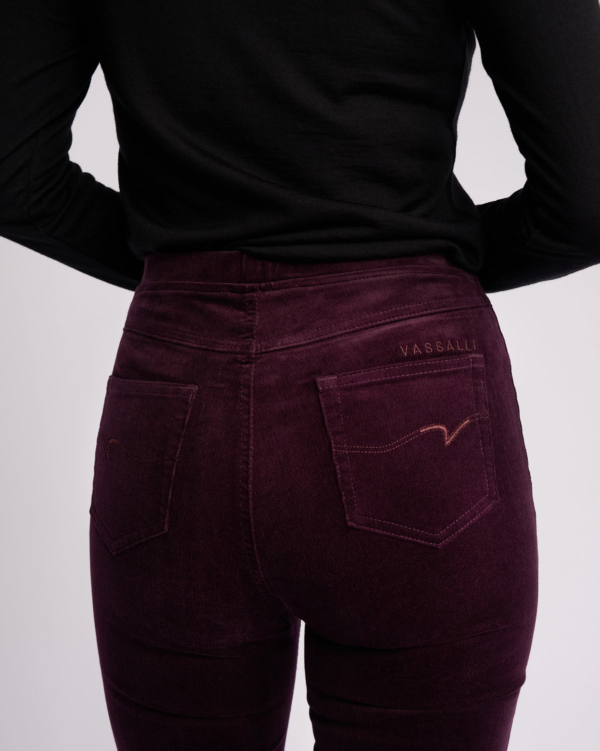 A person wearing a black long-sleeve top and Vassalli Bay Cord Pull On - Mulberry pants, shown from the waist down and back, highlighting the pocket details and stitching.
