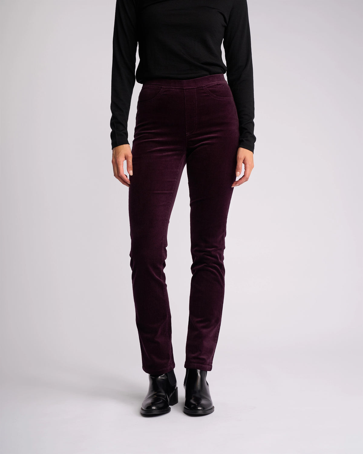 A person wearing a black long-sleeve top, Vassalli Bay Cord Pull On pants in Mulberry, and black ankle boots stands against a plain light background, facing forward with arms relaxed at their sides.