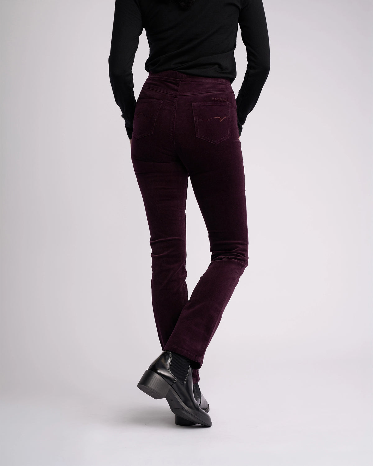 A person wearing a black long-sleeve top, black shoes, and Vassalli Bay Cord Pull On pants in mulberry stands with their back to the camera against a plain light background.