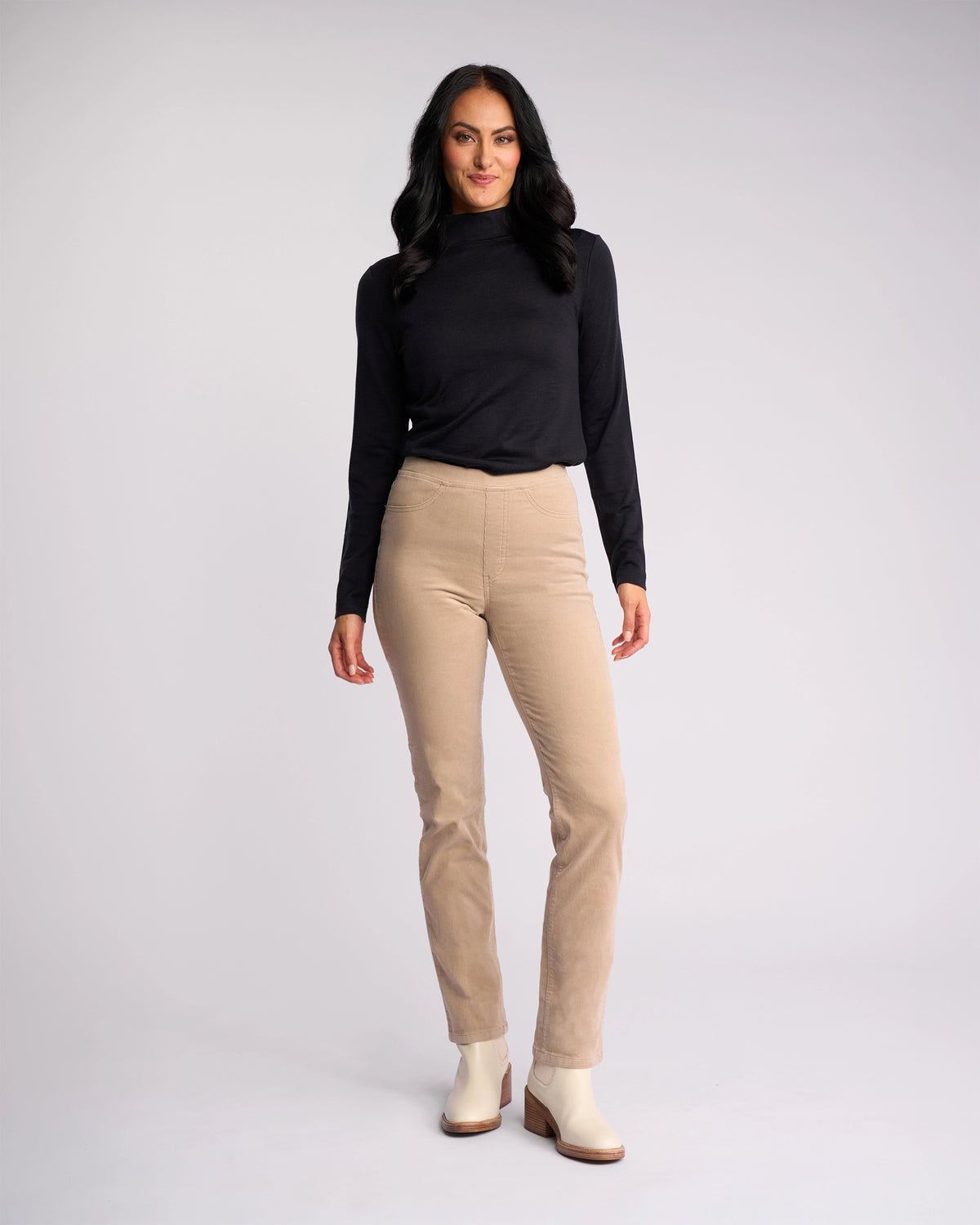 A woman with long dark hair, in a black long-sleeve top, Vassalli Bay Cord Pull On - Oat slim-leg pants, and light ankle boots, stands smiling against a plain light gray background.