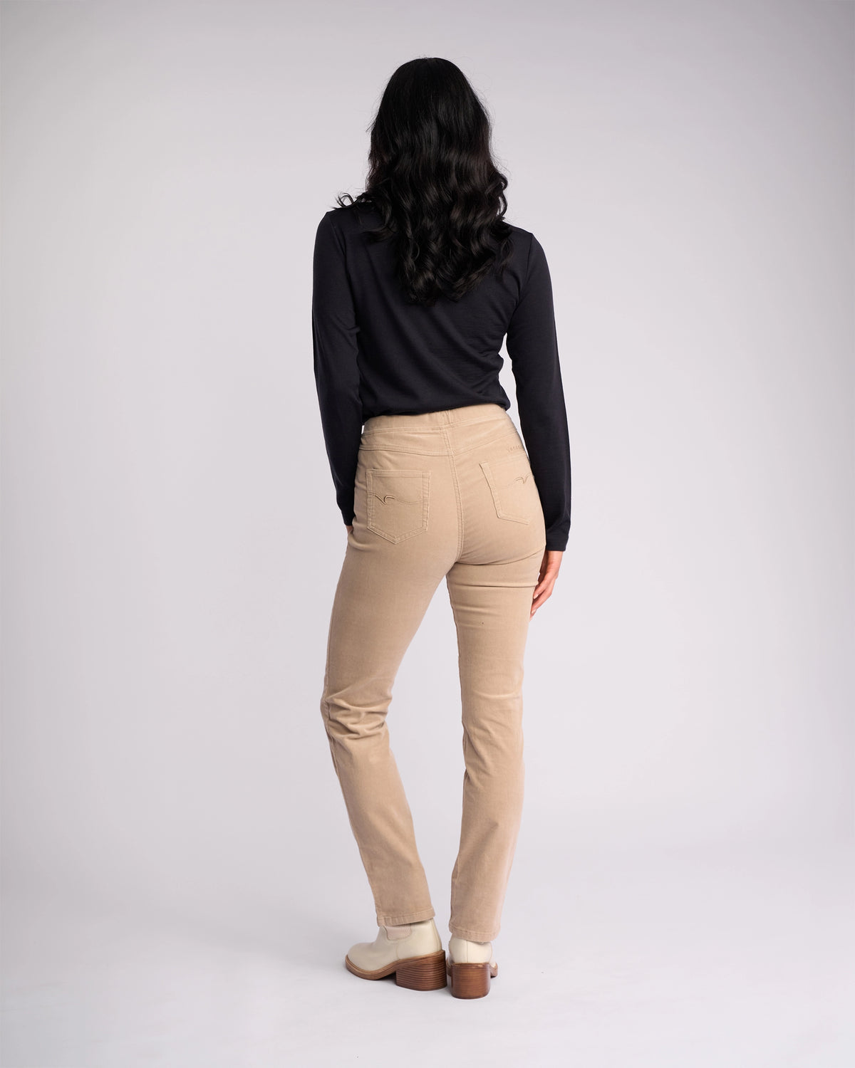 A woman faces away, wearing a Vassalli Bay Cord Pull On - Oat with a black long-sleeve top and beige shoes, her dark wavy hair down against a plain light background.
