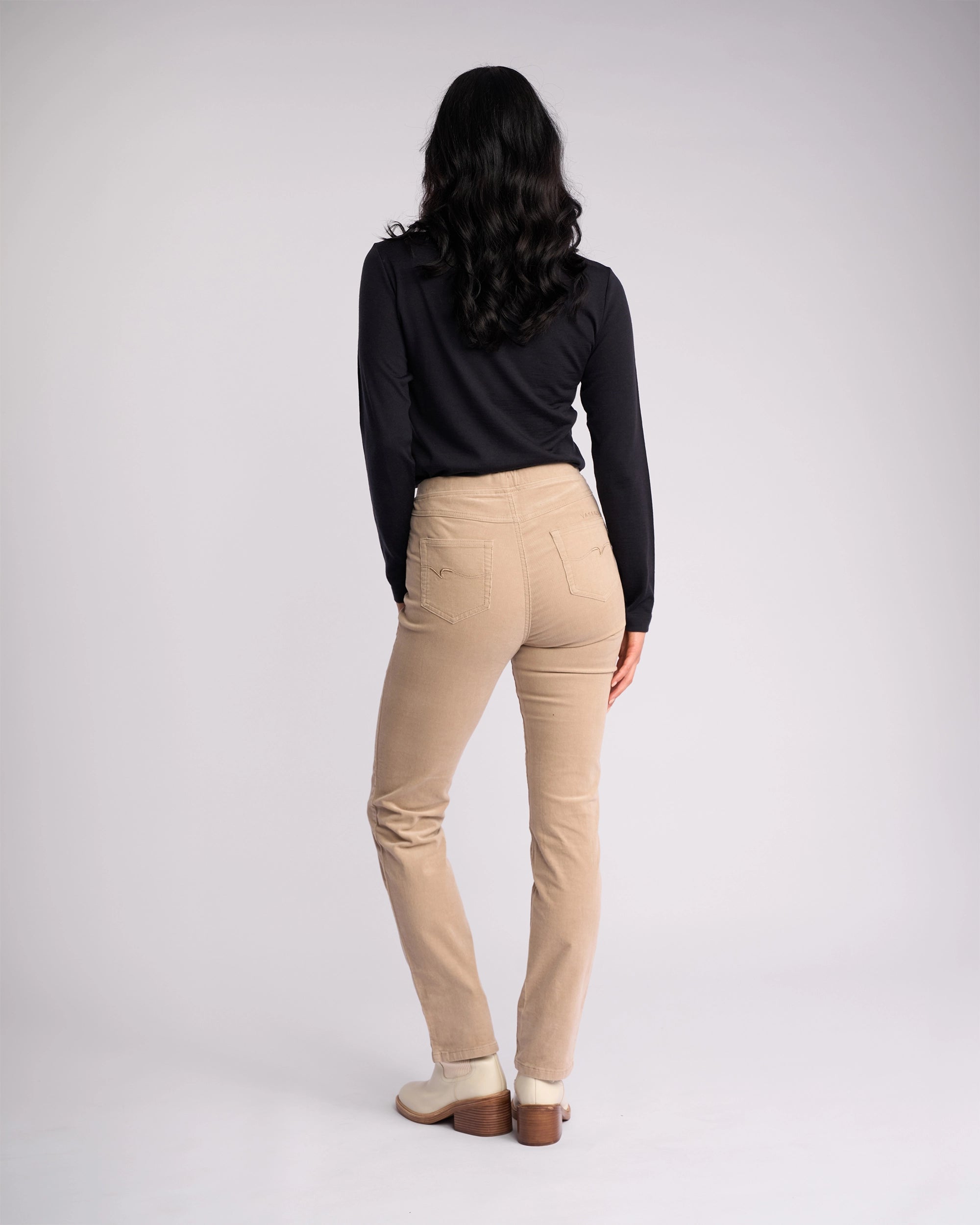 A woman faces away, wearing a Vassalli Bay Cord Pull On - Oat with a black long-sleeve top and beige shoes, her dark wavy hair down against a plain light background.