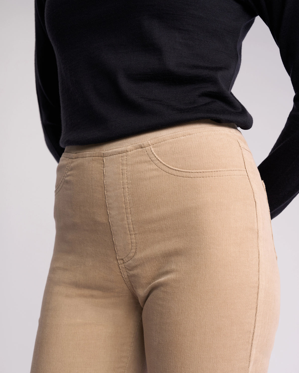 A person wearing a black long-sleeve top and Vassalli Bay Cord Pull On - Oat slim-leg pants stands with arms behind their back, shown from mid-torso to mid-thigh against a plain background.