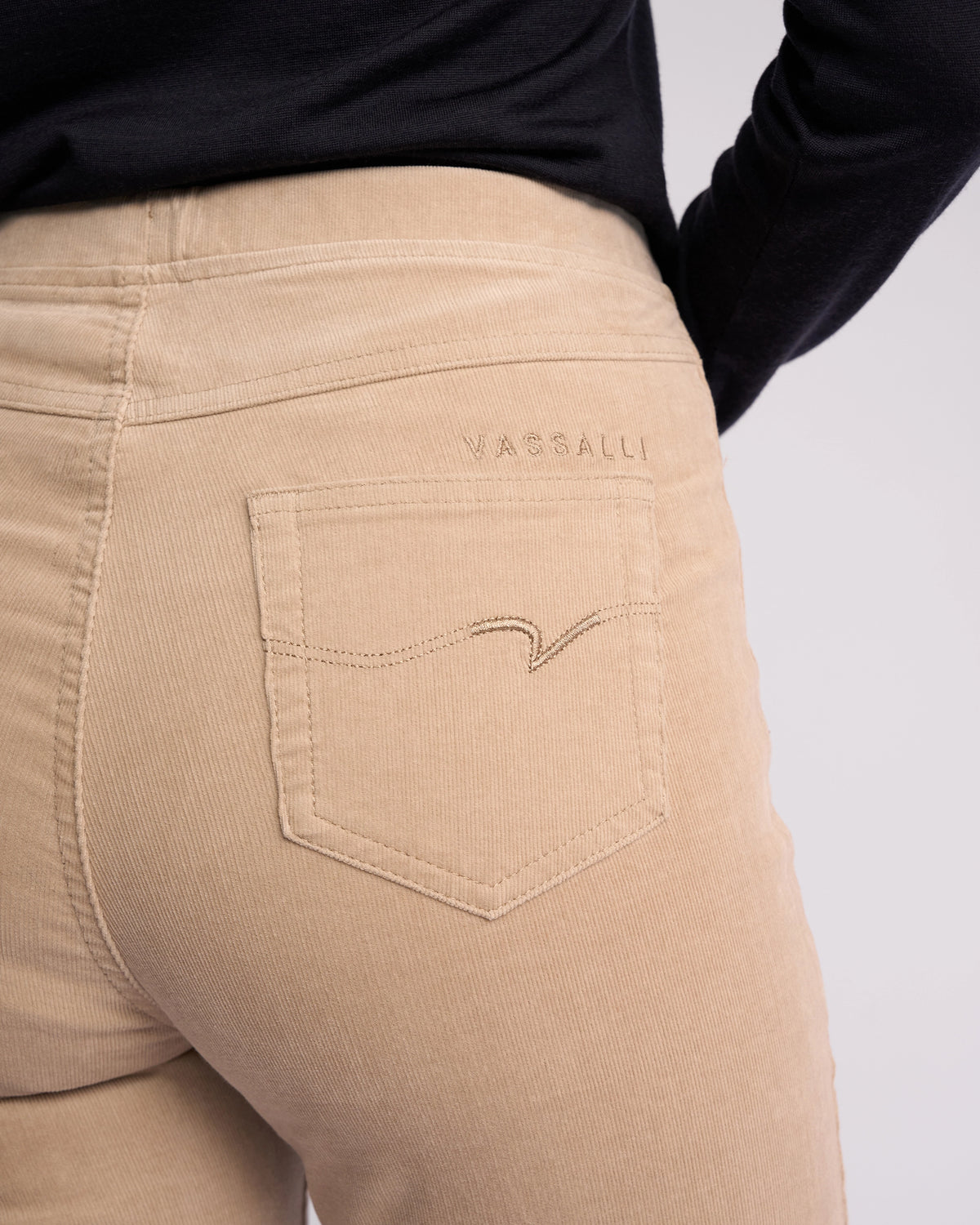 Close-up of a person wearing Vassalli Bay Cord Pull On pants in Oat, featuring a slim-leg fit, visible back pocket with stitched detailing, and "VASSALLI" embroidered above the pocket. The person pairs them with a black top.