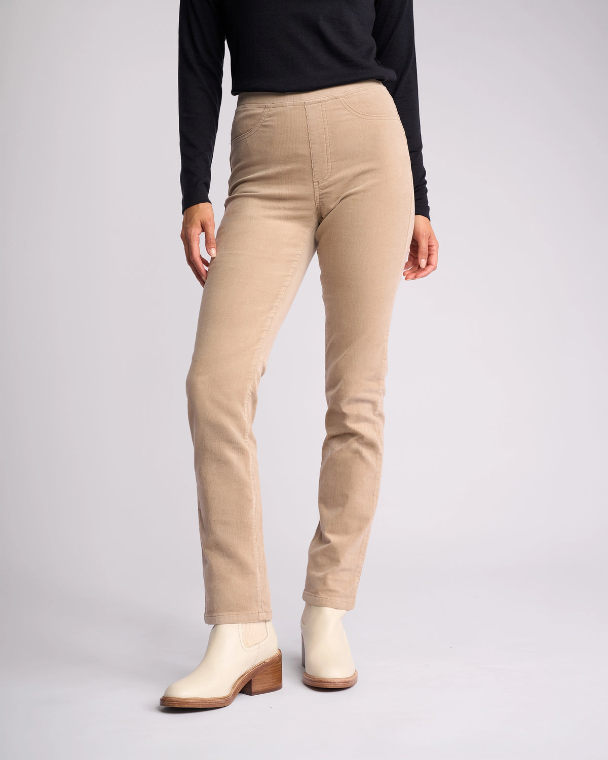 A person stands against a light gray background wearing a black long-sleeve top, Vassalli Bay Cord Pull On pants in Oat (beige, high-waisted, slim-leg), and cream ankle boots. The person's head is not visible.
