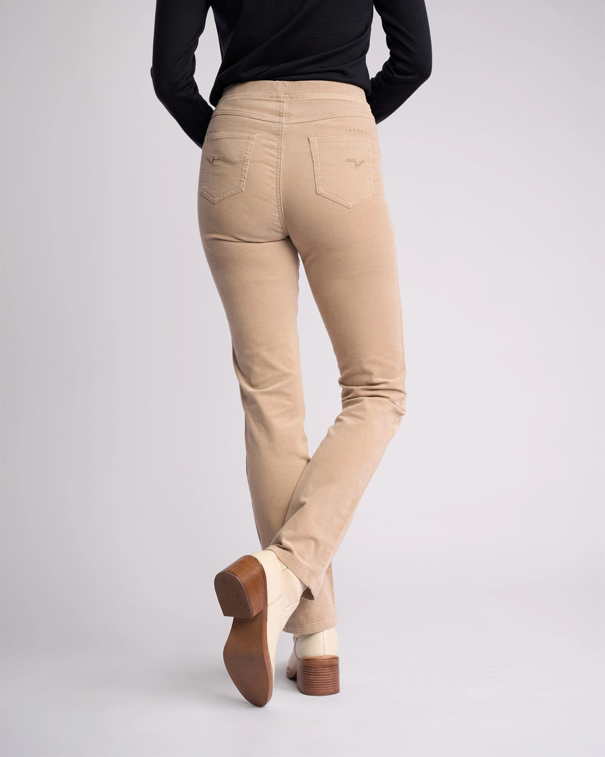 A person models the Vassalli Bay Cord Pull On - Oat pants with a black long-sleeve top, standing with one leg crossed behind the other to display the back view against a plain light background.