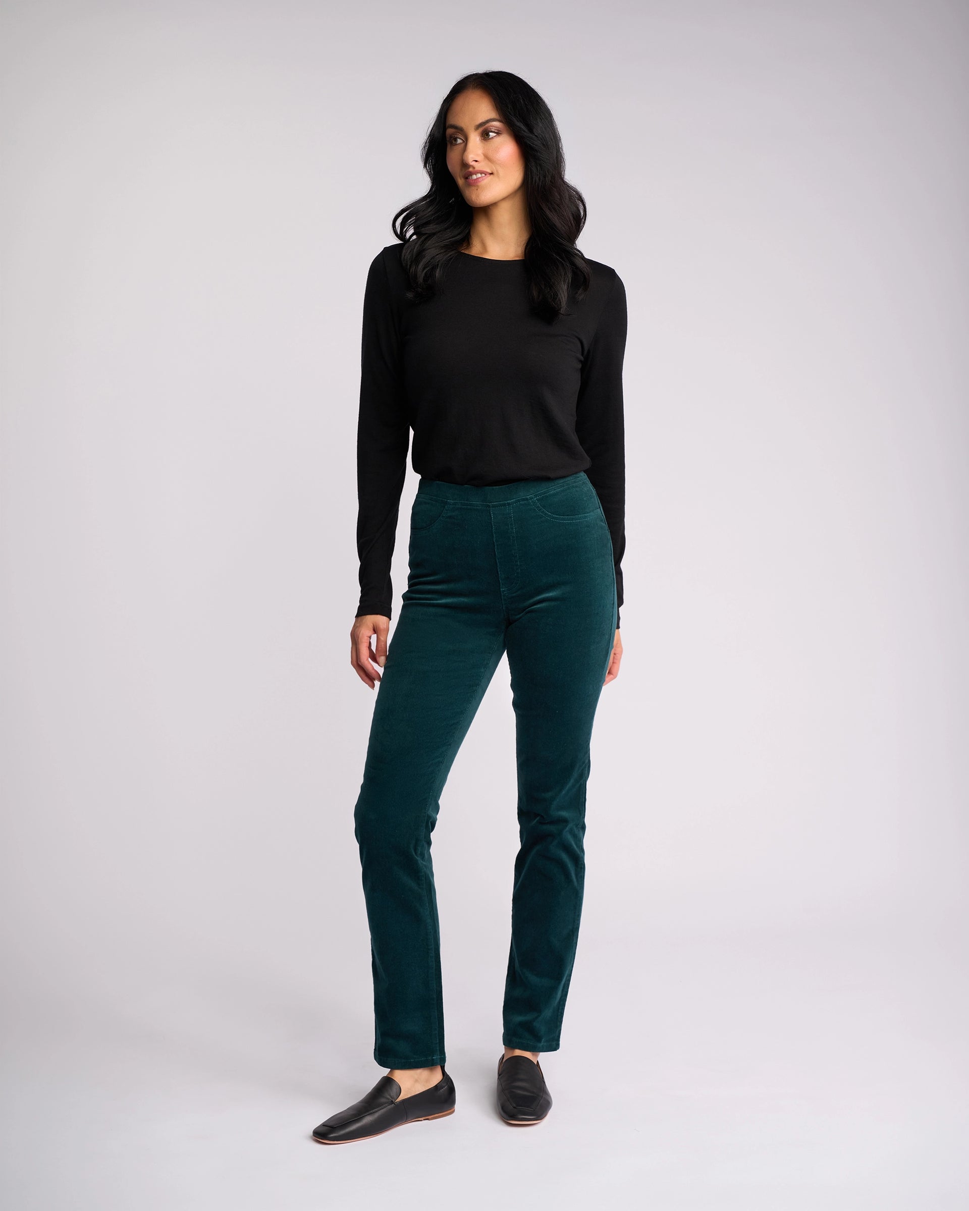 A woman with long dark hair wears a black long-sleeve top, Vassalli Bay Cord Pull On - Peacock slim-leg pants, and black slip-on shoes against a plain light gray background, looking slightly to the side.