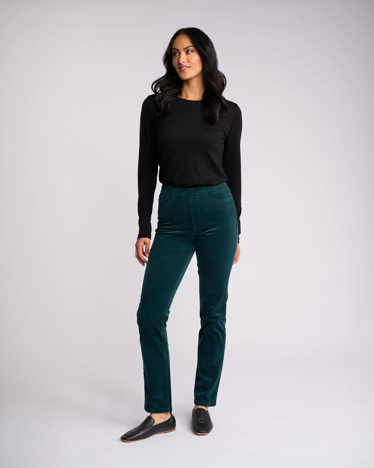 A woman with long dark hair wears a black long-sleeve top, Vassalli Bay Cord Pull On - Peacock slim-leg pants, and black slip-on shoes against a plain light gray background, looking slightly to the side.