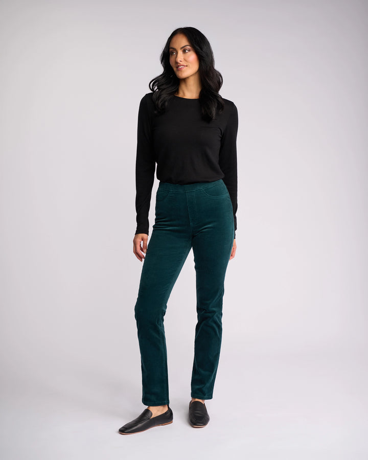 A woman with long dark hair wears a black long-sleeve top, Vassalli Bay Cord Pull On - Peacock slim-leg pants, and black slip-on shoes against a plain light gray background, looking slightly to the side.