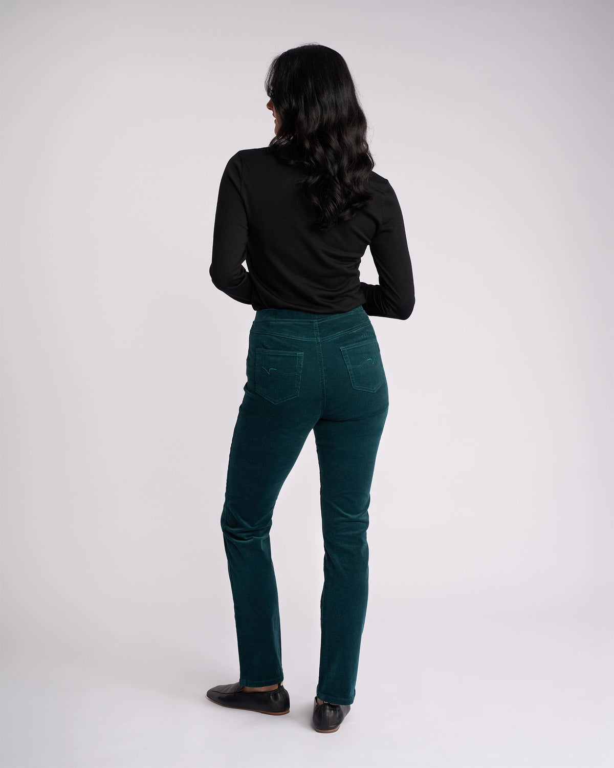 A woman with long dark hair, wearing a black long-sleeve top and Vassalli’s Bay Cord Pull On - Peacock pants, stands facing away from the camera against a plain light background.