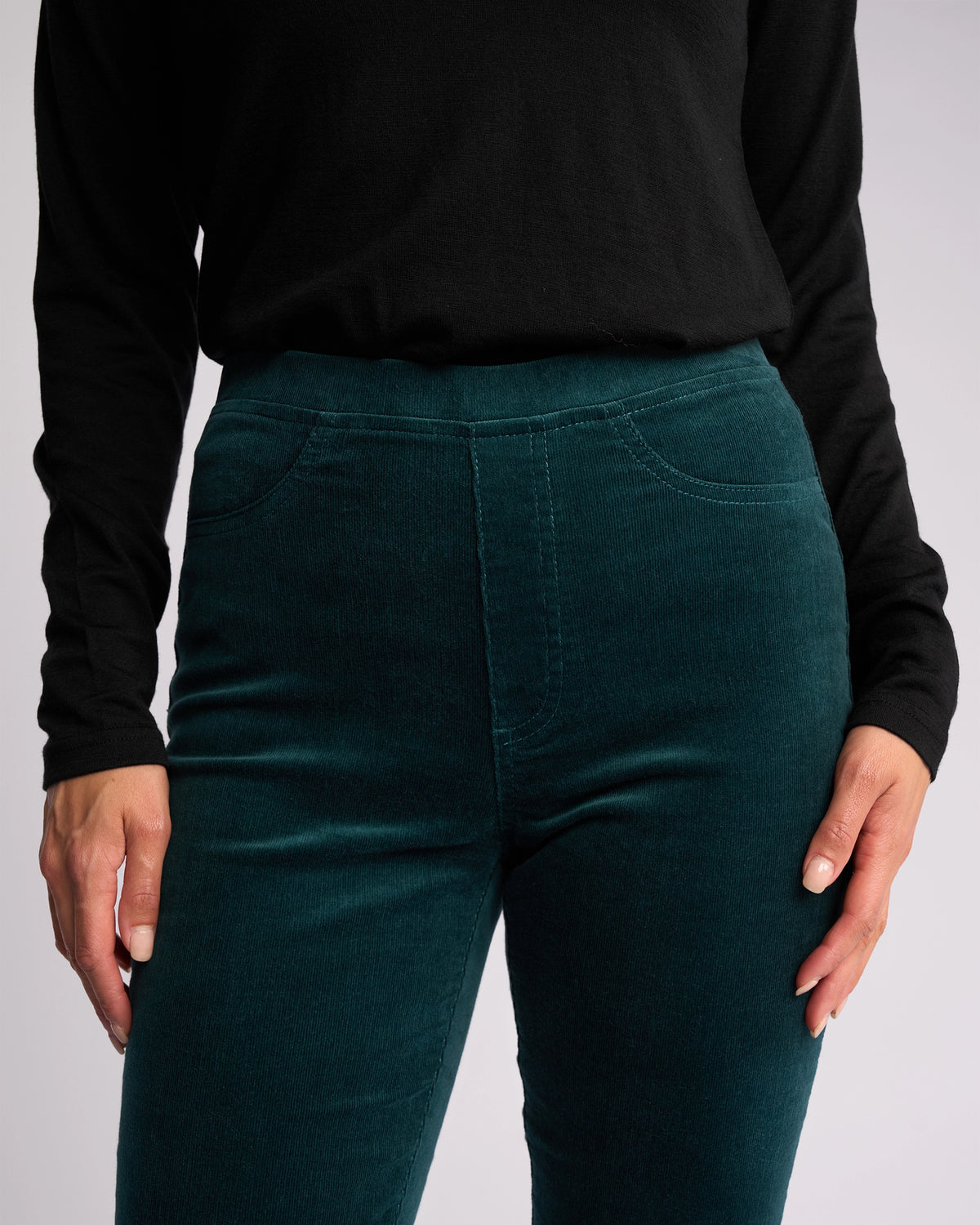 A person stands with hands relaxed at their sides, wearing a black long-sleeve top and Vassalli's Bay Cord Pull On - Peacock high-waisted, dark green stretch pinwale cord pants.