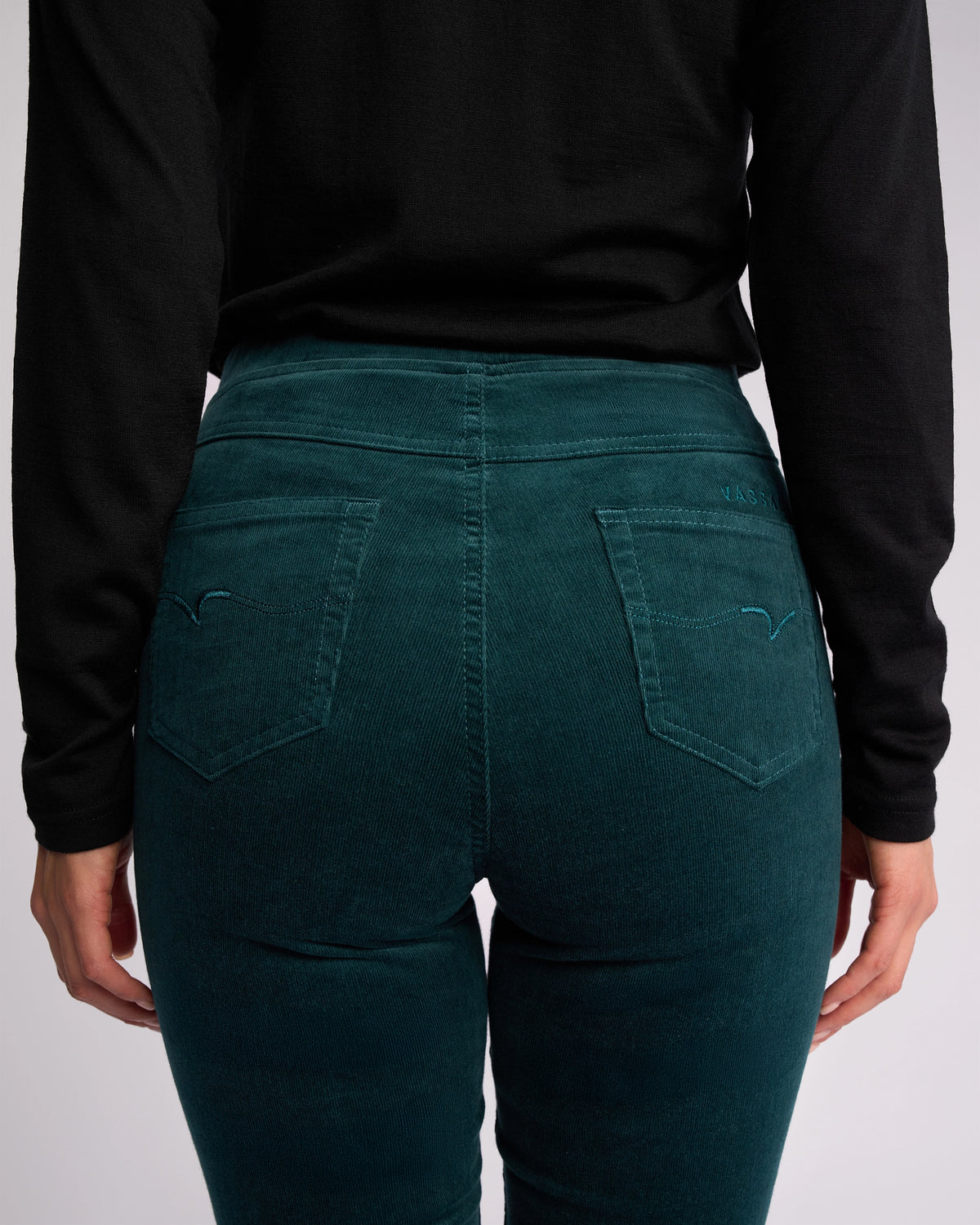 A person is shown from the back wearing a black long-sleeve top and Vassalli Bay Cord Pull On - Peacock slim-leg pants, with focus on the waistband and back pockets.