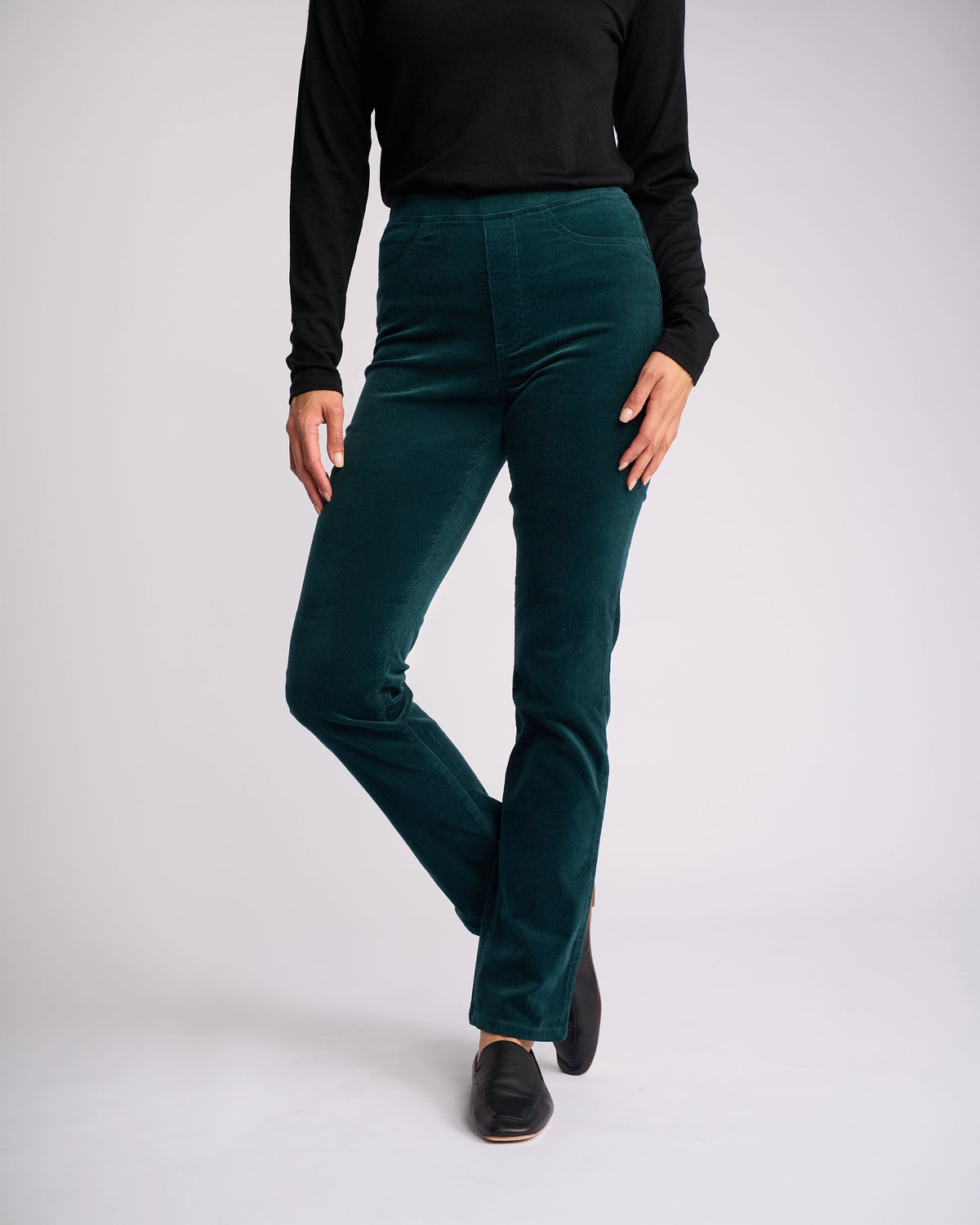 A person stands against a plain light background, wearing a black long-sleeve top, Vassalli Bay Cord Pull On pants in Peacock (dark green) with a slim-leg fit, and black slip-on shoes. One leg is slightly bent; their face is not visible.