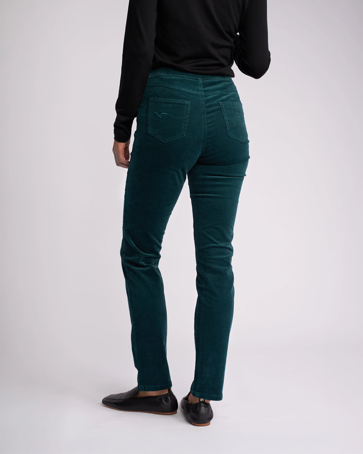 A person wearing Vassalli Bay Cord Pull On pants in Peacock, a black long-sleeve top, and black shoes stands with their back to the camera against a plain, light background.