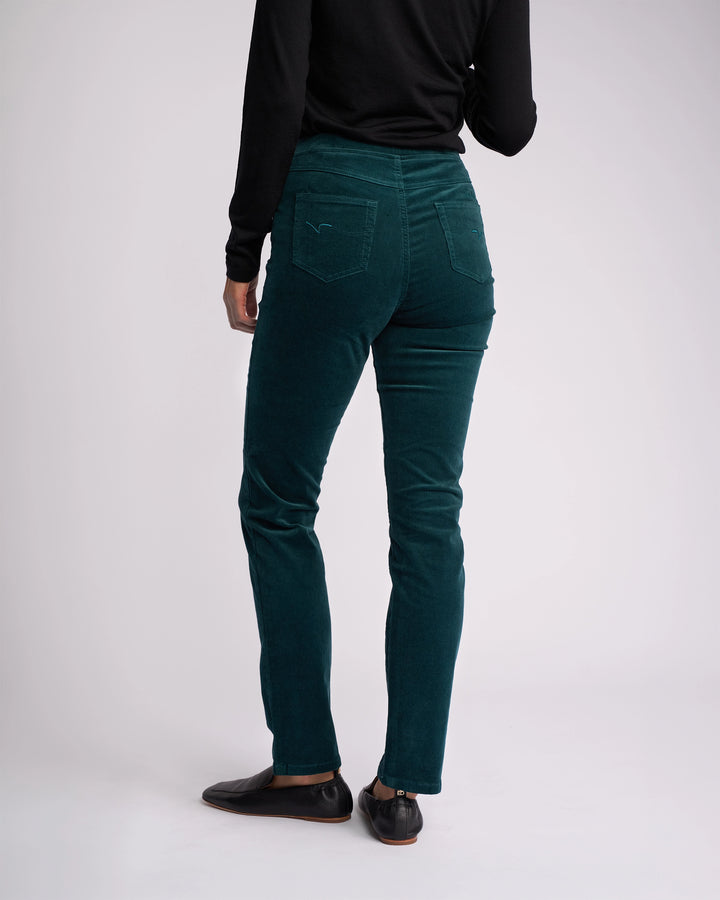 A person wearing Vassalli Bay Cord Pull On pants in Peacock, a black long-sleeve top, and black shoes stands with their back to the camera against a plain, light background.