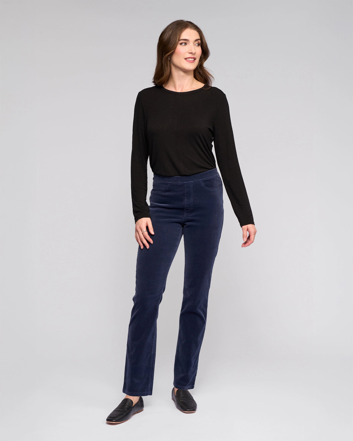 A woman stands against a light gray background, smiling and wearing Vassalli's Bay Cord Pull On - Slate pants with a long-sleeve black top and black shoes. Her arms are relaxed at her sides as she looks slightly to the side.