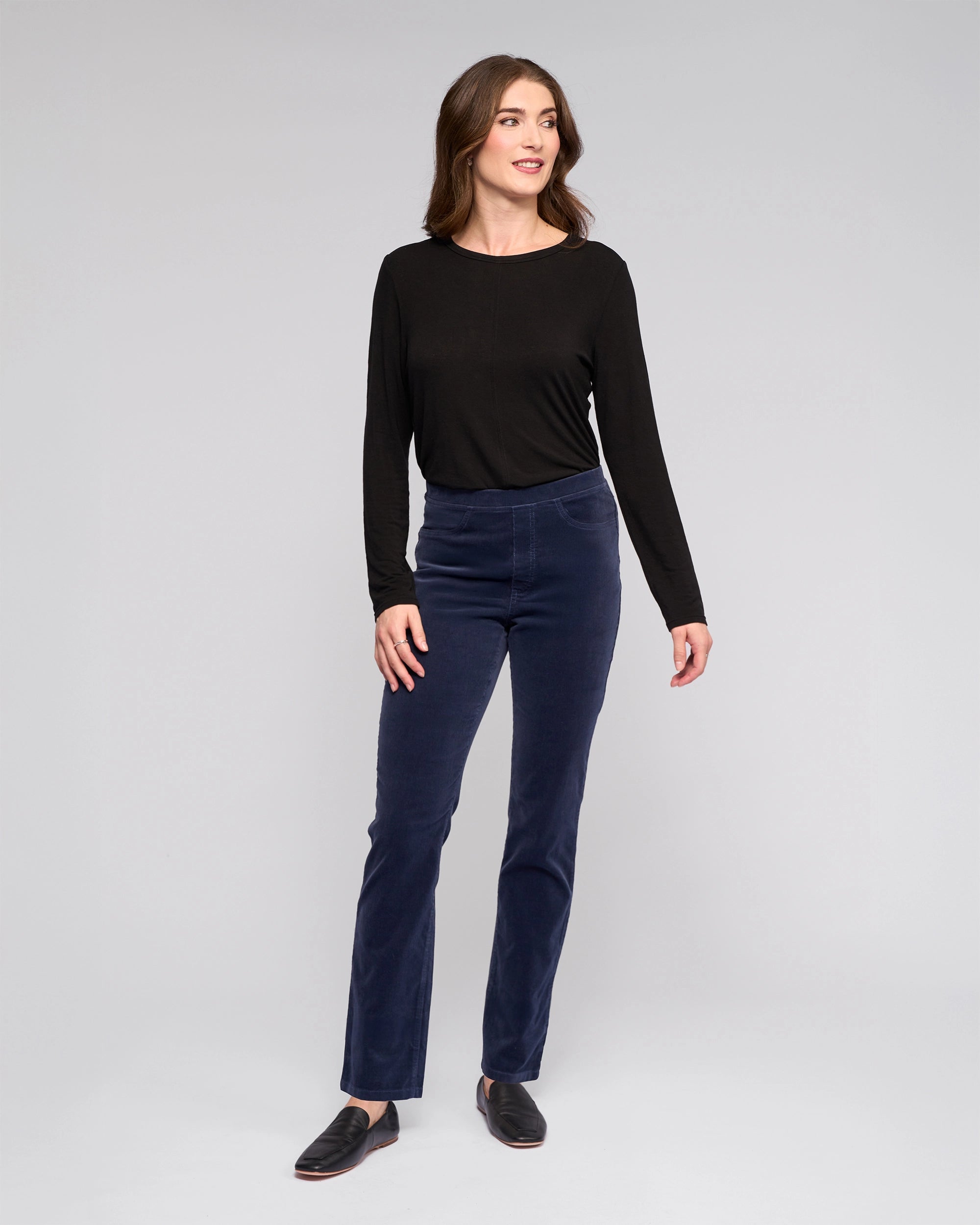 A woman stands against a light gray background, smiling and wearing Vassalli's Bay Cord Pull On - Slate pants with a long-sleeve black top and black shoes. Her arms are relaxed at her sides as she looks slightly to the side.