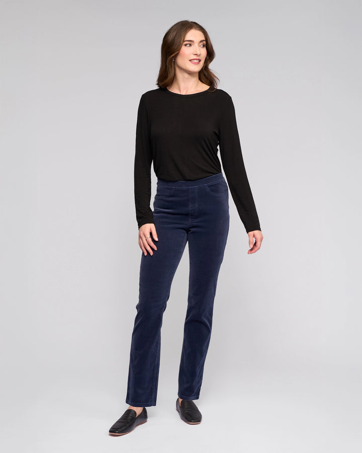 A woman stands against a light gray background, smiling and wearing Vassalli's Bay Cord Pull On - Slate pants with a long-sleeve black top and black shoes. Her arms are relaxed at her sides as she looks slightly to the side.