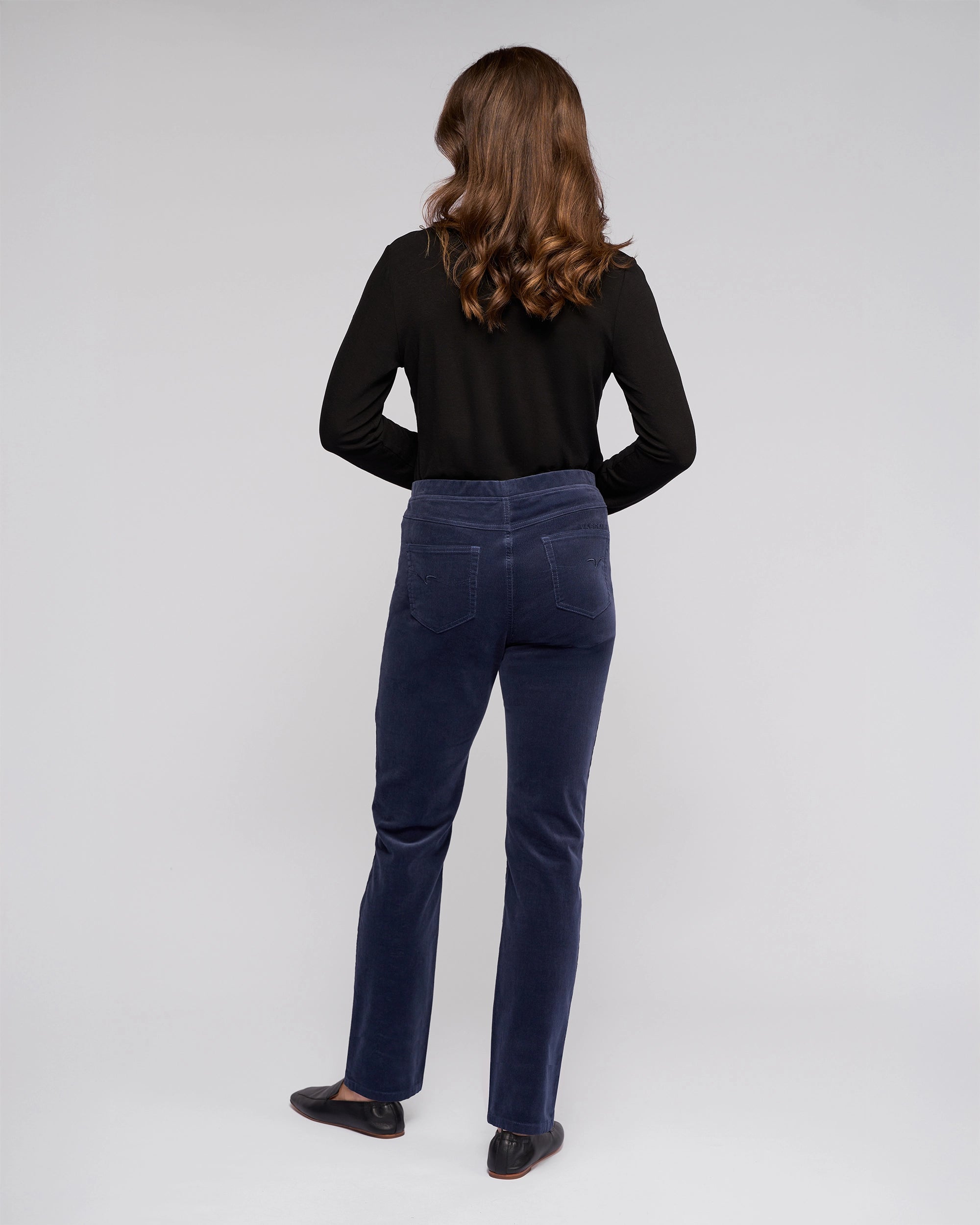 A person with long brown hair, dressed in a black long-sleeve top, Vassalli Bay Cord Pull On - Slate pants, and black shoes, stands facing away against a plain light background.