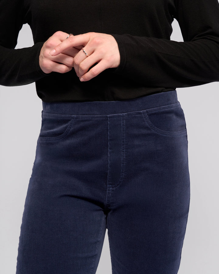 A person wears Vassalli's Bay Cord Pull On - Slate, a dark blue high-rise slim-leg pant, with a black long-sleeve top and a silver ring. Their hands are clasped, and the image is cropped from shoulders to mid-thigh.