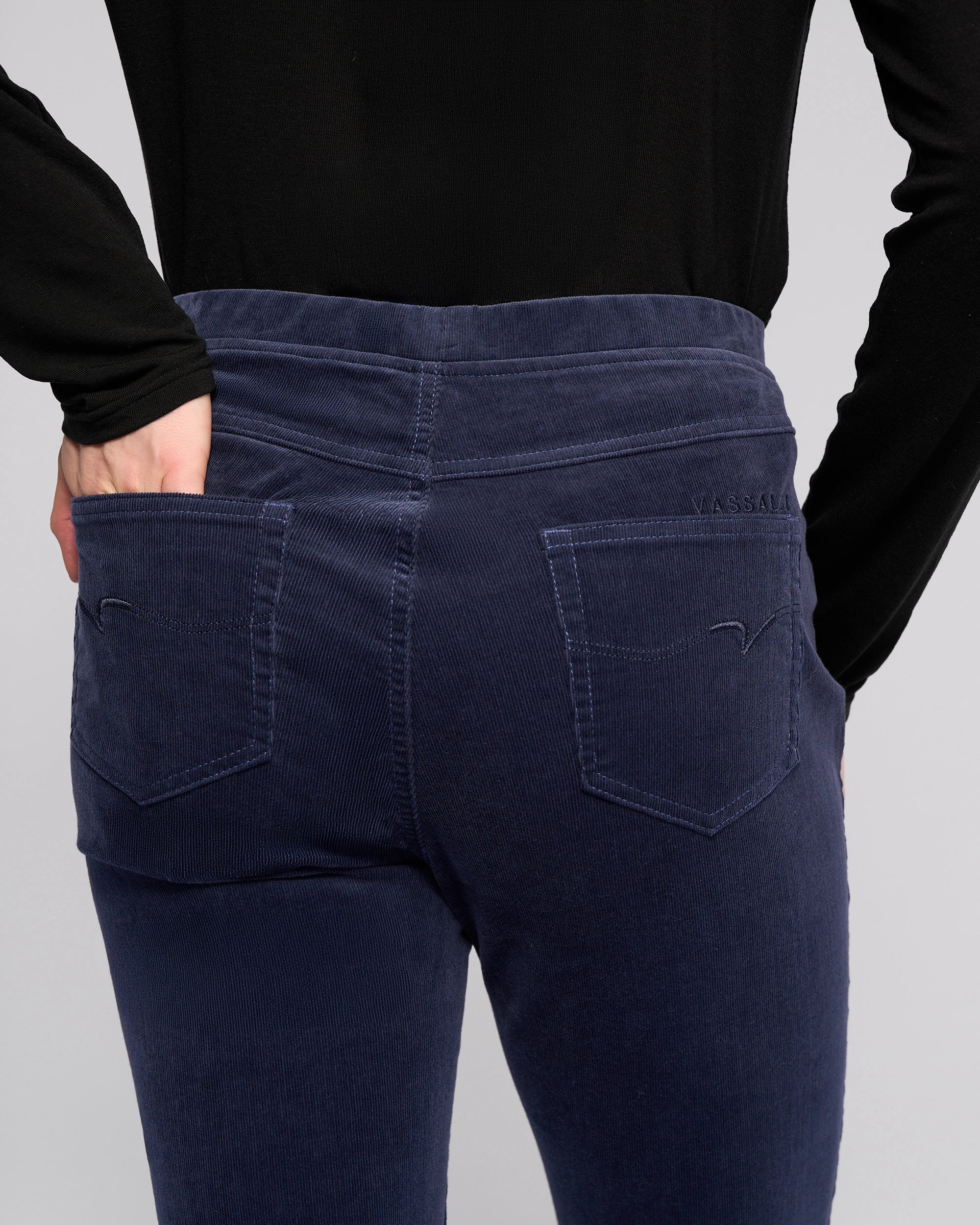 A person wearing a black long-sleeve top and Vassalli Bay Cord Pull On pants in Slate, seen from the back with one hand in a back pocket.