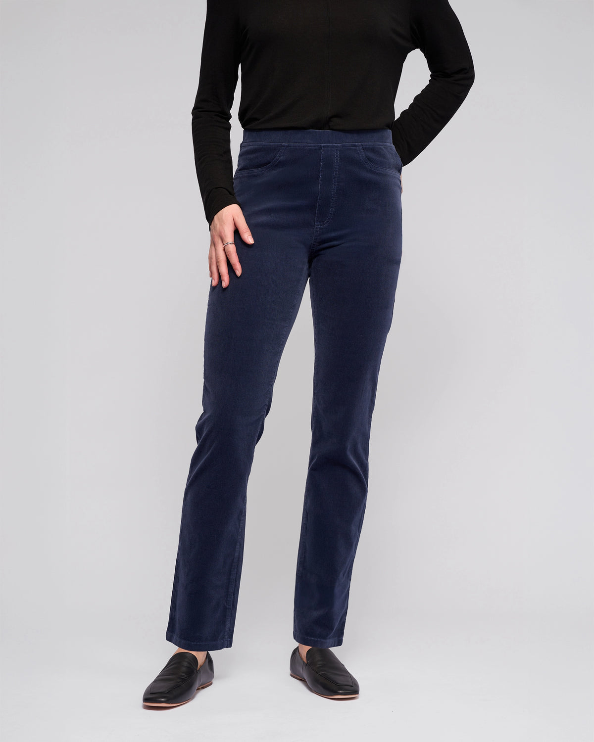 A person wears a long-sleeved black top, Vassalli Bay Cord Pull On - Slate pants, and black slip-on shoes against a plain light gray background. Their head is not visible in the image.