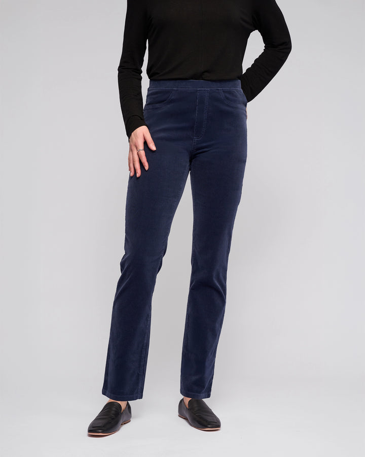 A person wears a long-sleeved black top, Vassalli Bay Cord Pull On - Slate pants, and black slip-on shoes against a plain light gray background. Their head is not visible in the image.