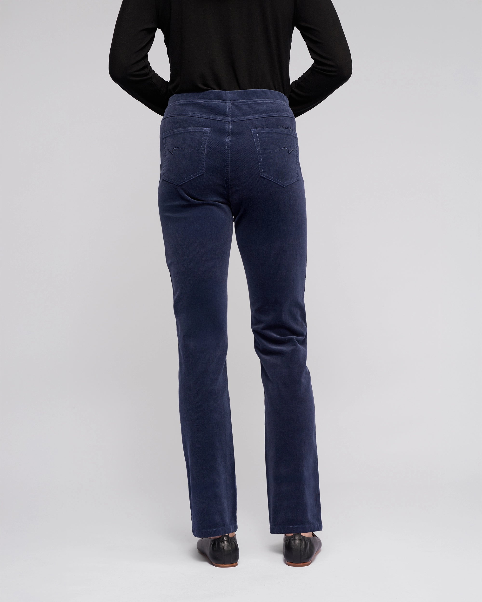 A person wearing a long-sleeve black top and Vassalli Bay Cord Pull On pants in Slate stands facing away from the camera, displaying the back of the outfit with black shoes against a plain light background.