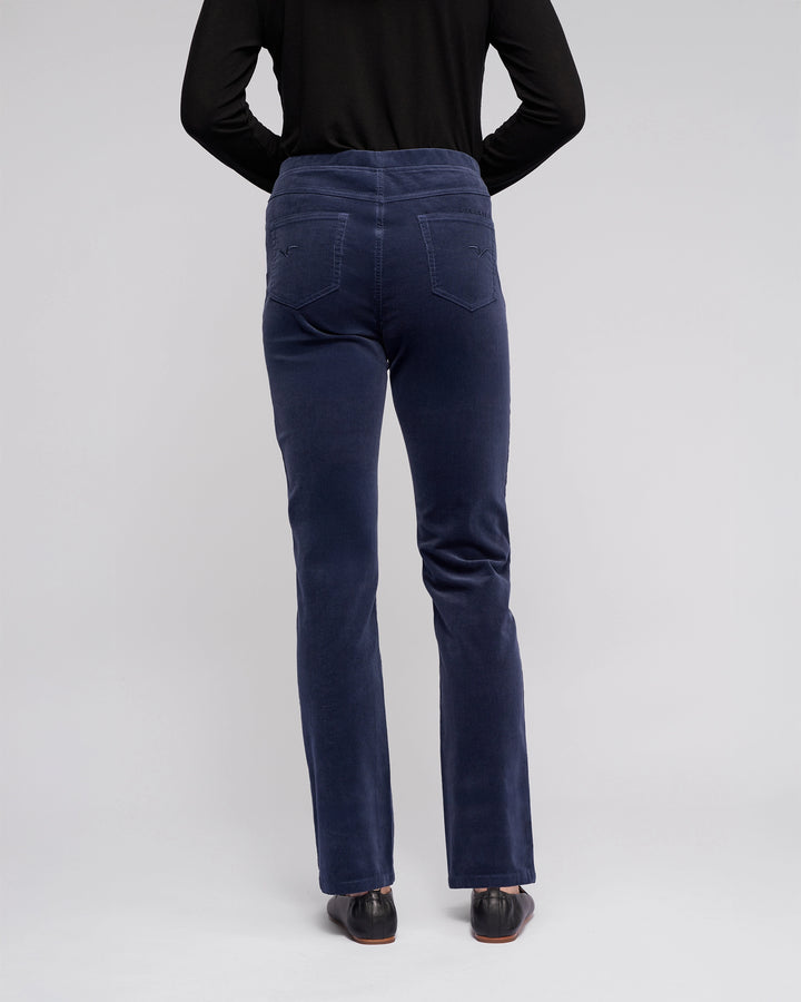 A person wearing a long-sleeve black top and Vassalli Bay Cord Pull On pants in Slate stands facing away from the camera, displaying the back of the outfit with black shoes against a plain light background.