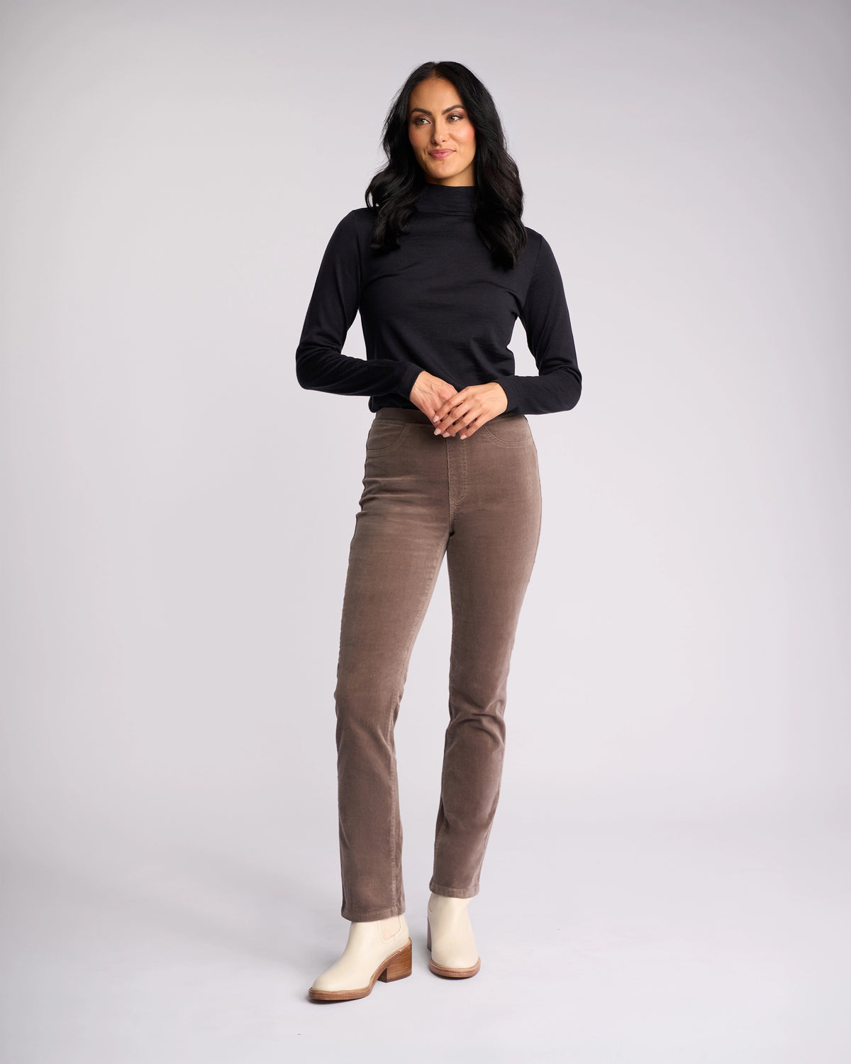 A woman with long dark hair stands against a light background, wearing the Vassalli Bay Cord Pull On - Taupe pants paired with a black long-sleeve top and cream ankle boots, smiling slightly with one hand on her hip.