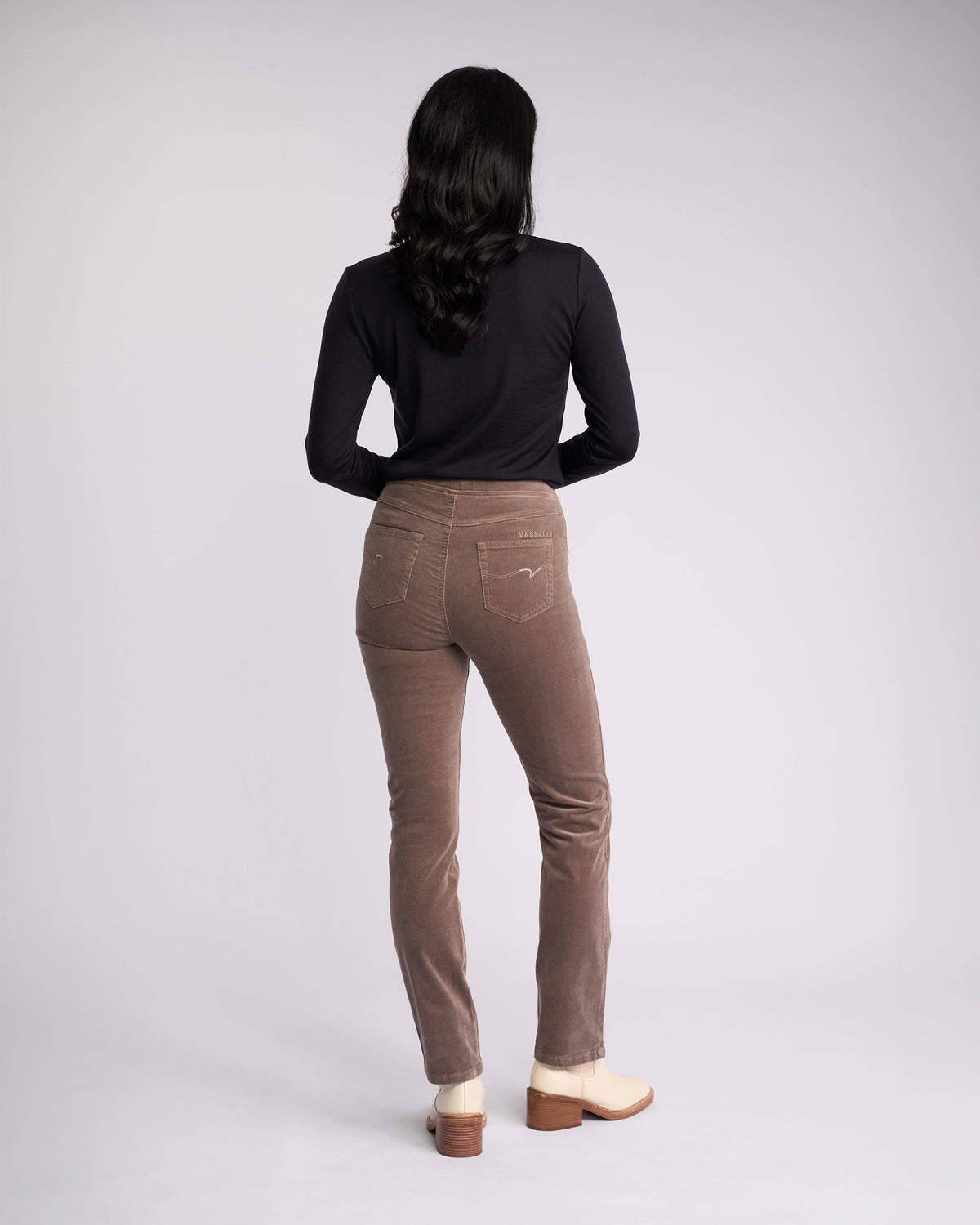 A person with long dark hair, wearing a black long-sleeve top and Vassalli Bay Cord Pull On pants in taupe, stands facing away from the camera against a plain light background.