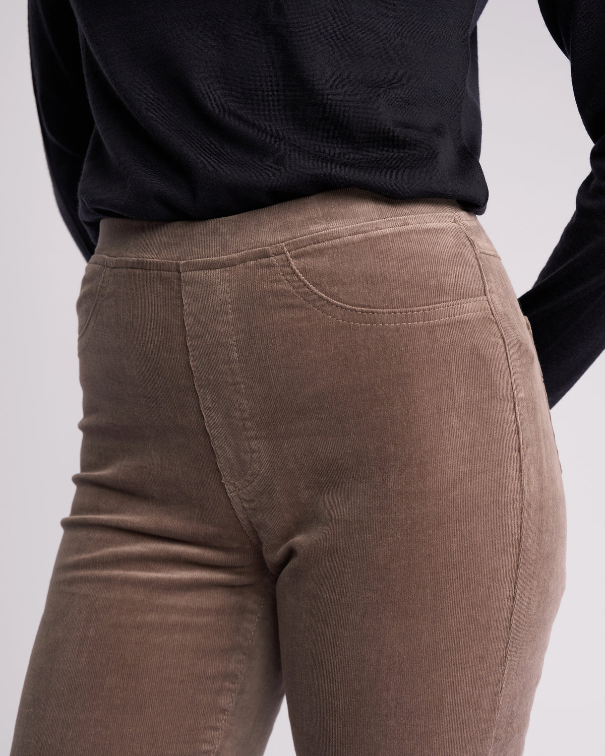 A person wears the Vassalli Bay Cord Pull On in taupe—high-waisted, light brown stretch cord pants—paired with a black long-sleeve top, shown from waist to mid-thigh against a neutral background.